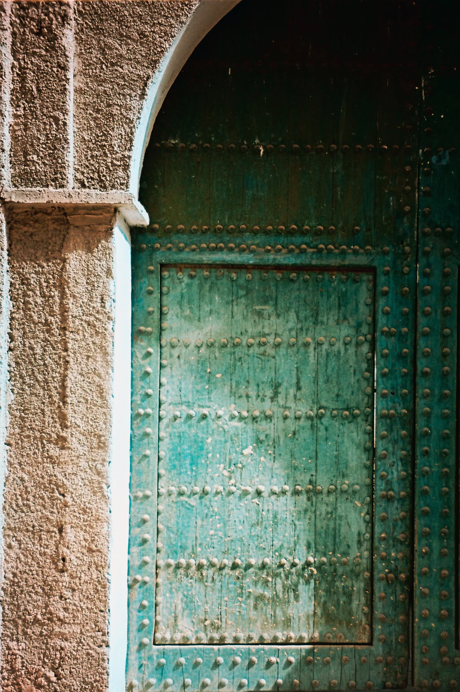Close-up of an old green wooden door with metal studs, set in a textured stone wall with an arch above.