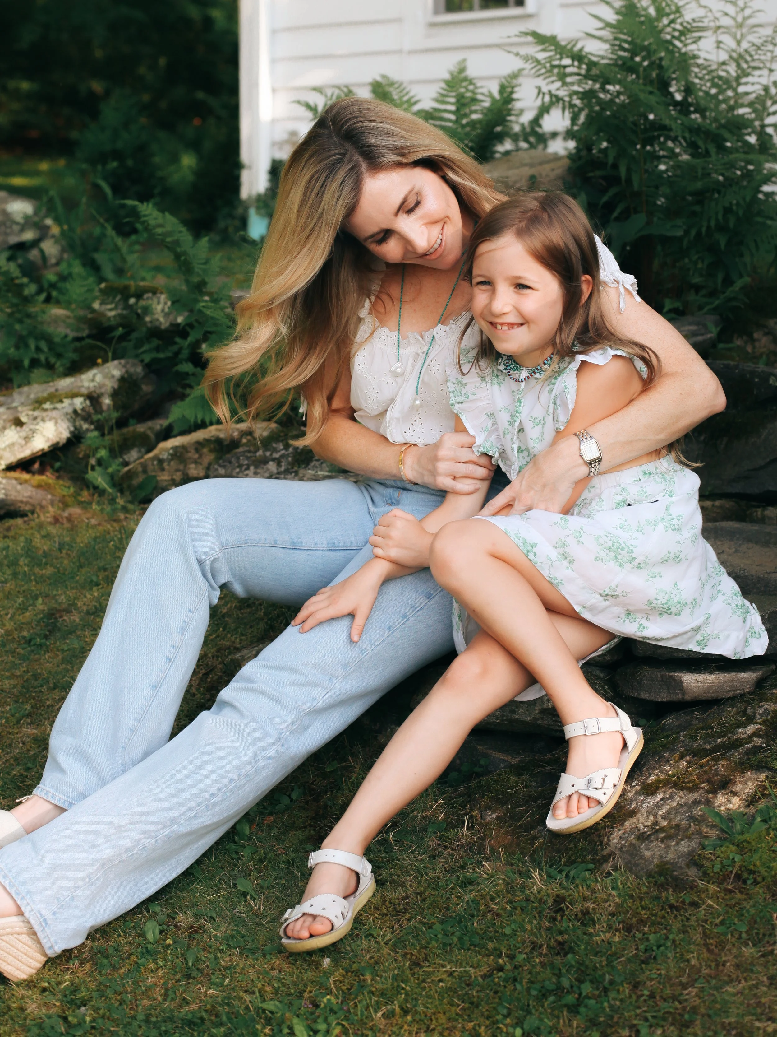 A woman and young girl sitting on rocks outdoors, hugging and smiling. The woman has long, wavy blonde hair and is wearing a white blouse and light blue jeans. The girl has long brown hair and is wearing a white dress with green patterns and white sa