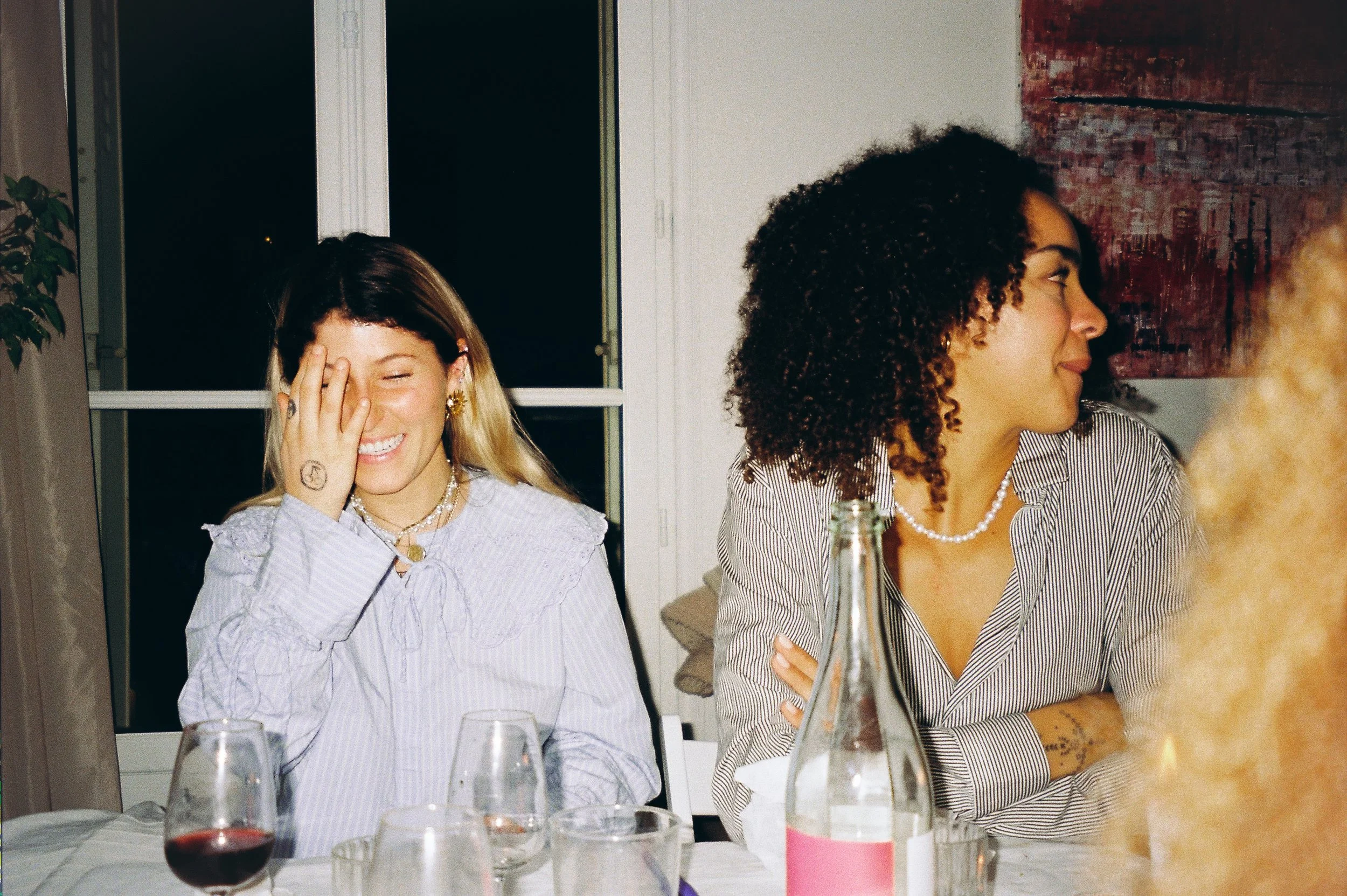 Two women sitting at a table during a dinner, one is laughing with her hand covering part of her face, the other is looking to her side with a smile, with a bottle and glasses on the table.