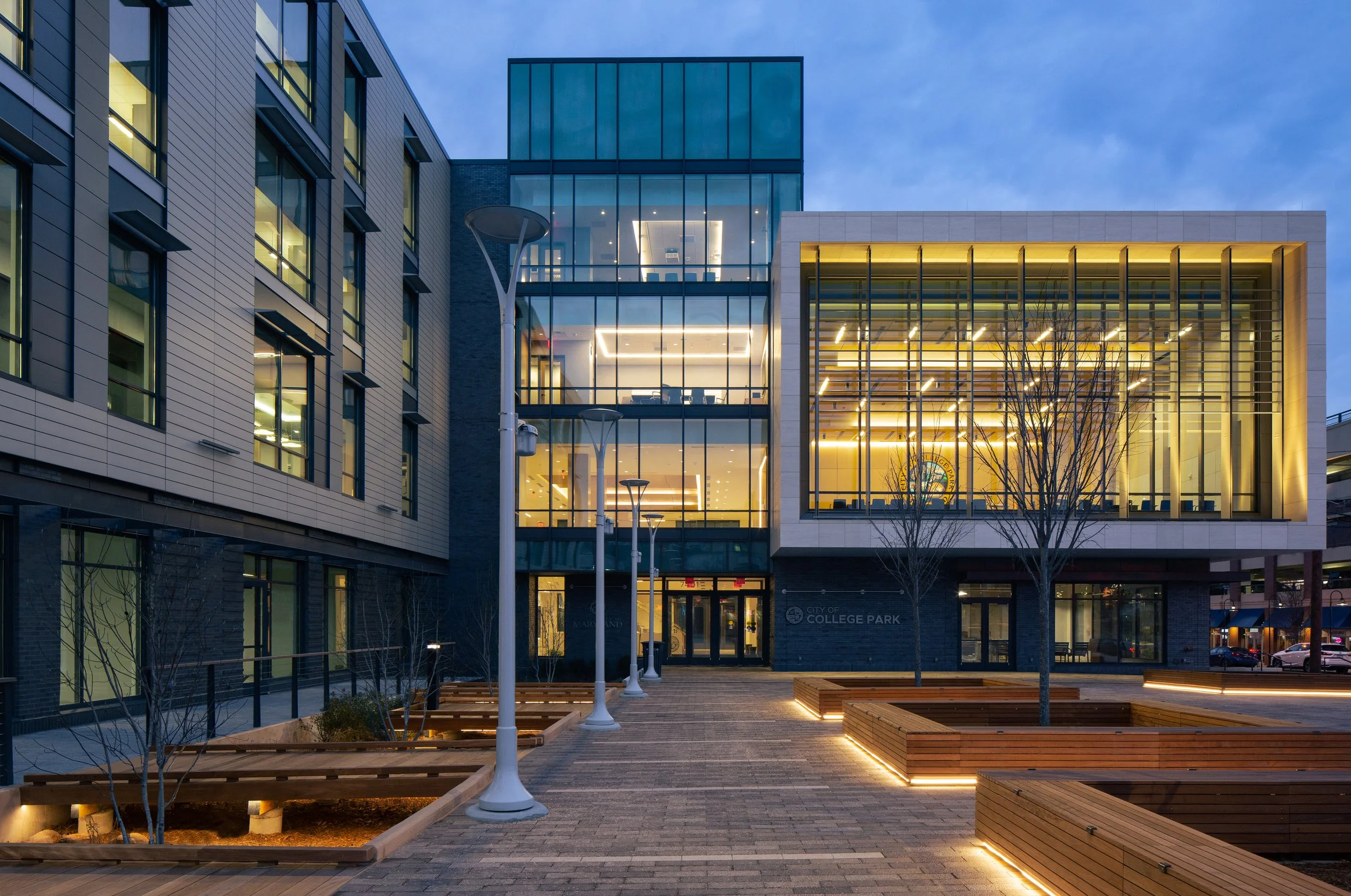 Twilight / Sunset shot of the newly constructed College Park City Hall. The sky is a deep blue, the lights within the windows of the building are glowing a warm amber. There is landscape lighting around the plants and benches. Architecture.