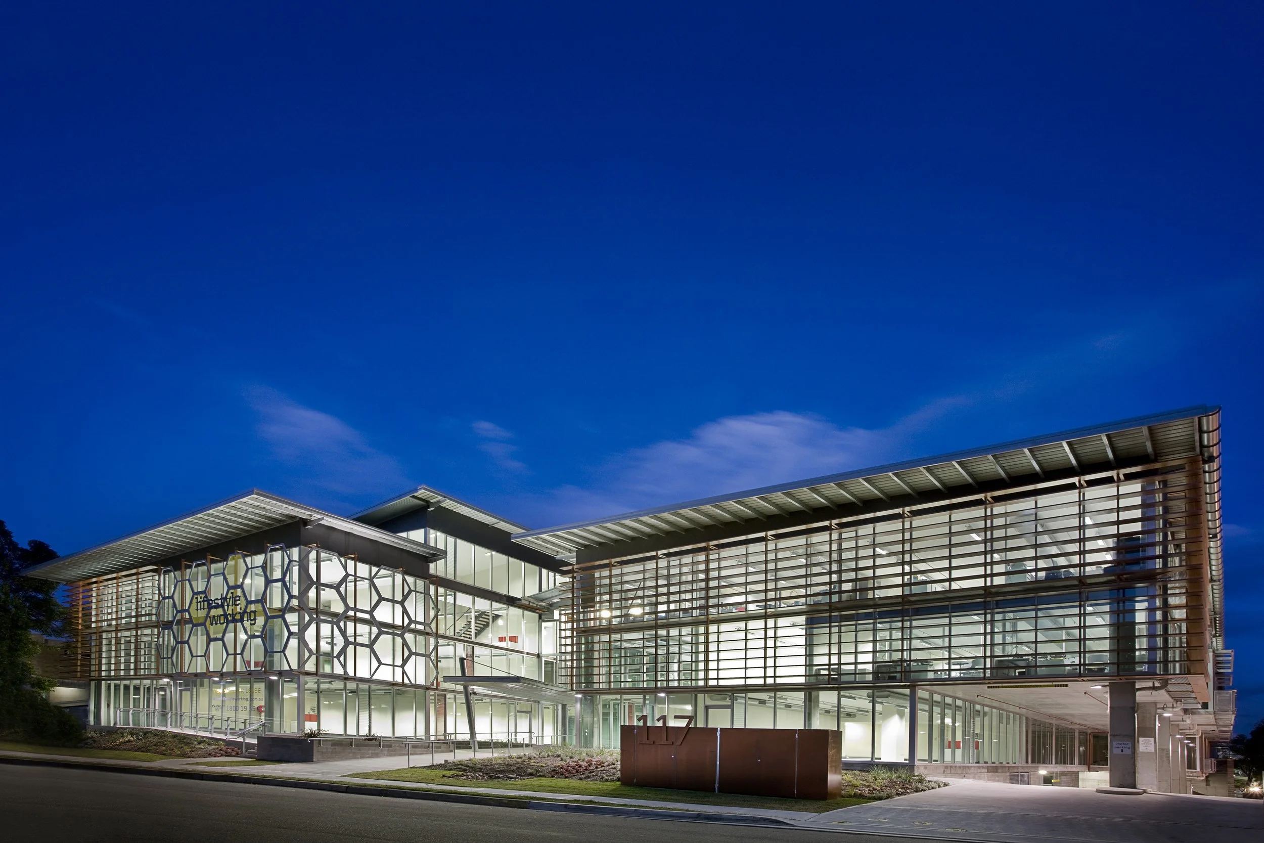 Nighttime exterior of a modern, two-story building with glass walls and a geometric honeycomb pattern on part of the facade. The building is well-lit with interior lights visible through the windows.