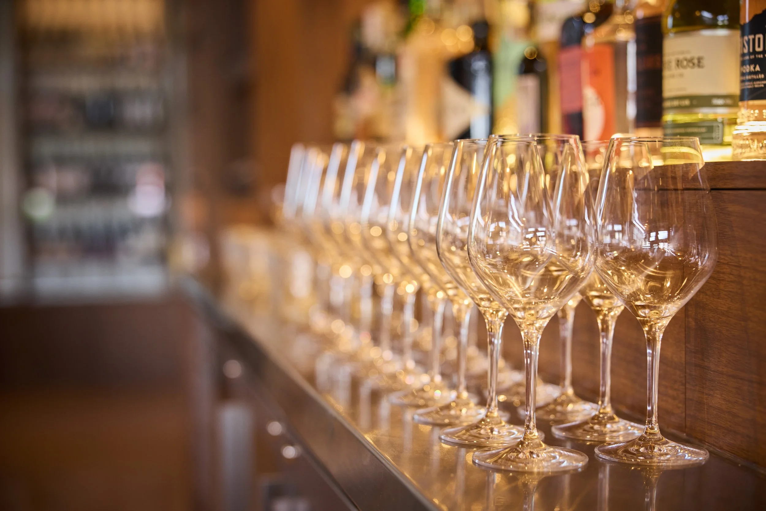 A row of empty wine glasses on a bar counter with bottles of alcohol on a shelf behind.