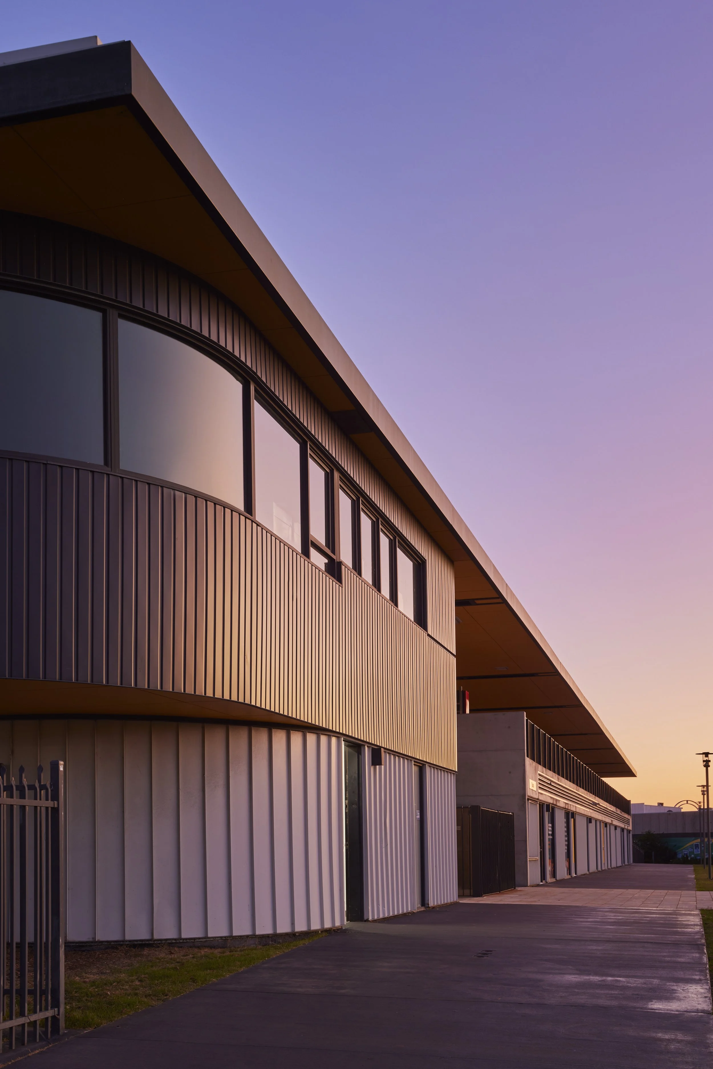 Modern building with curved and straight metal siding, large windows, and a sidewalk at sunset.