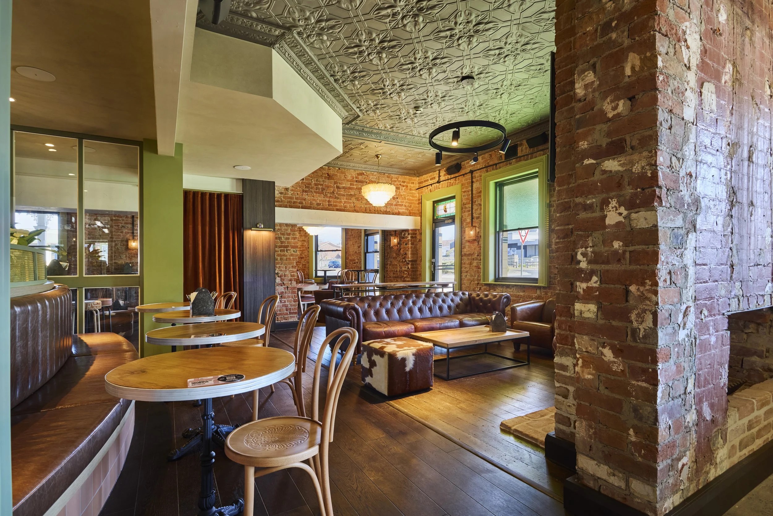 Interior of a cozy cafe with exposed brick walls, green window frames, wooden flooring, and vintage furniture including leather sofas and wooden chairs, illuminated by natural light from large windows.