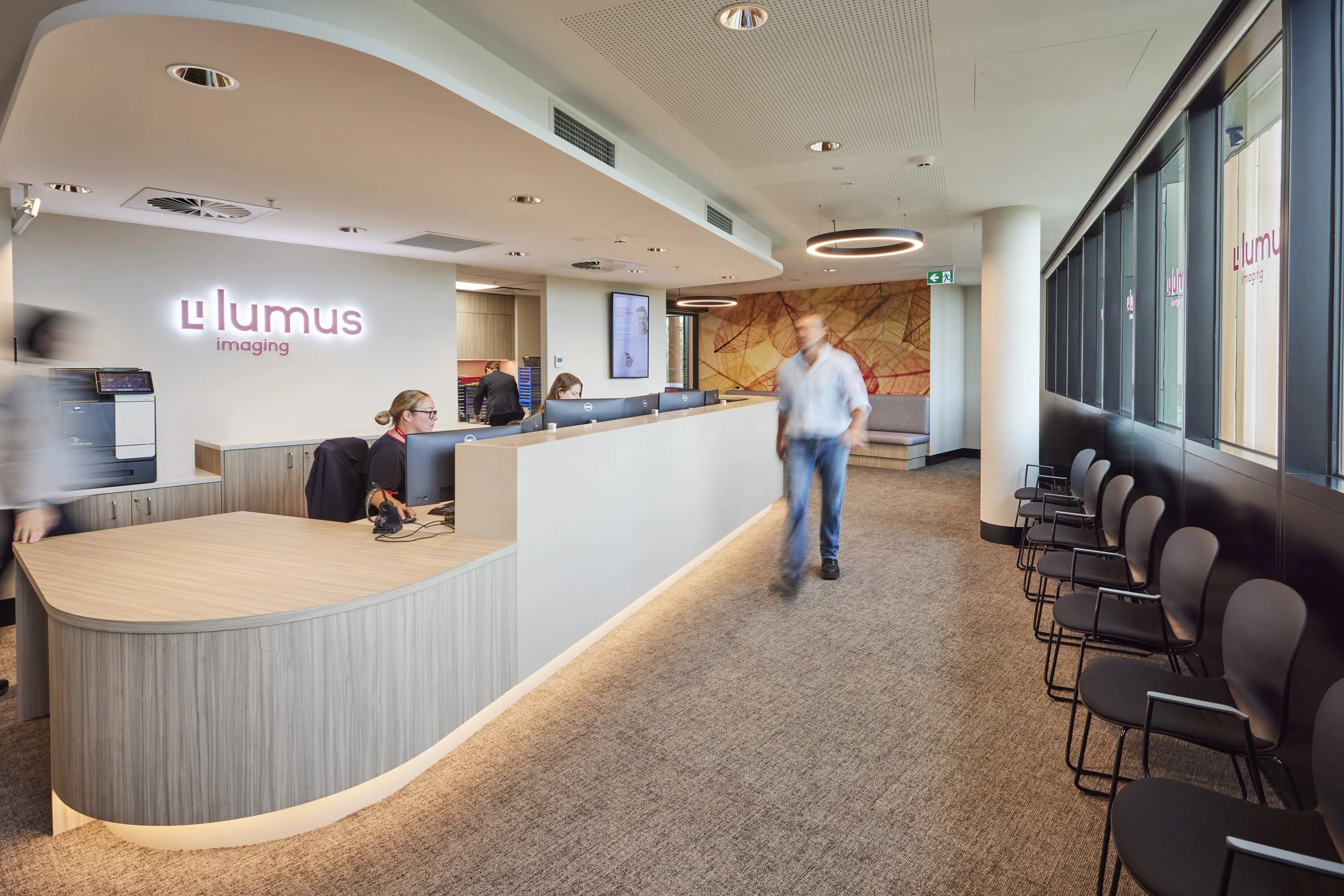 Lobby area of Lumus Imaging featuring a reception desk with staff working on computers, a row of black chairs, a wall with the Lumus logo, and people walking in the space.