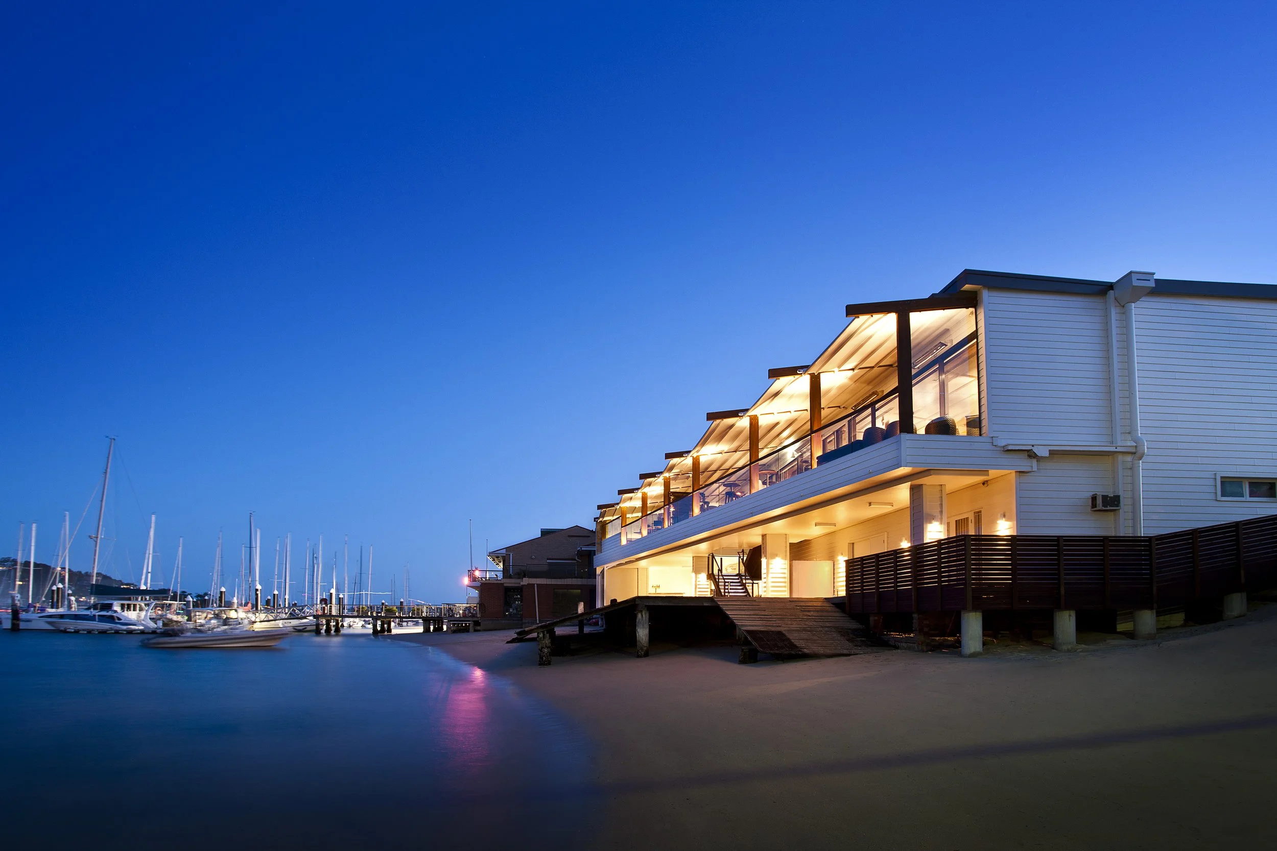 A modern waterfront house with large glass windows and outdoor lighting at dusk, situated near a marina with boats and sailboats docked.