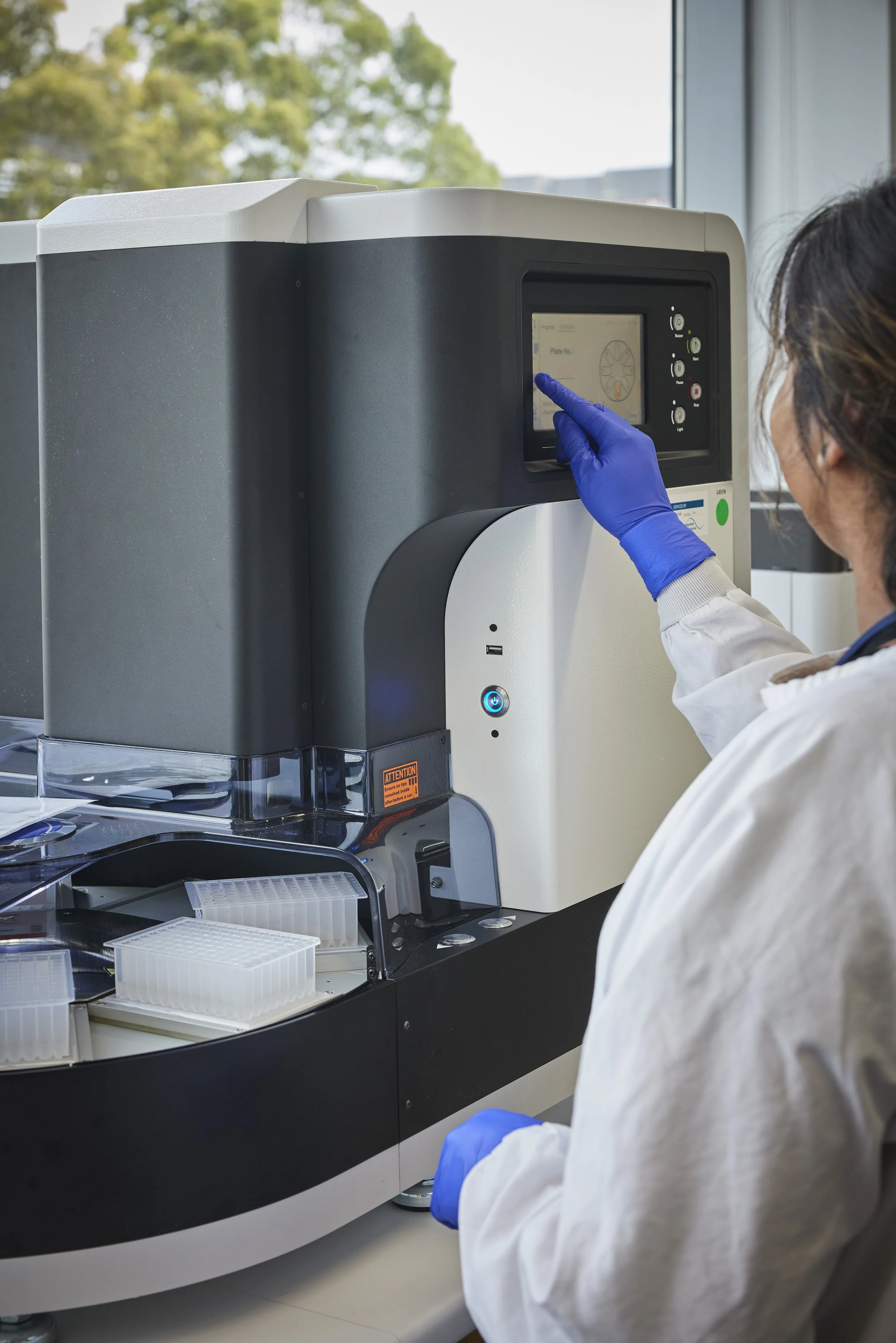 A scientist in a lab coat and blue gloves operating a large scientific machine with a touchscreen in a laboratory setting.