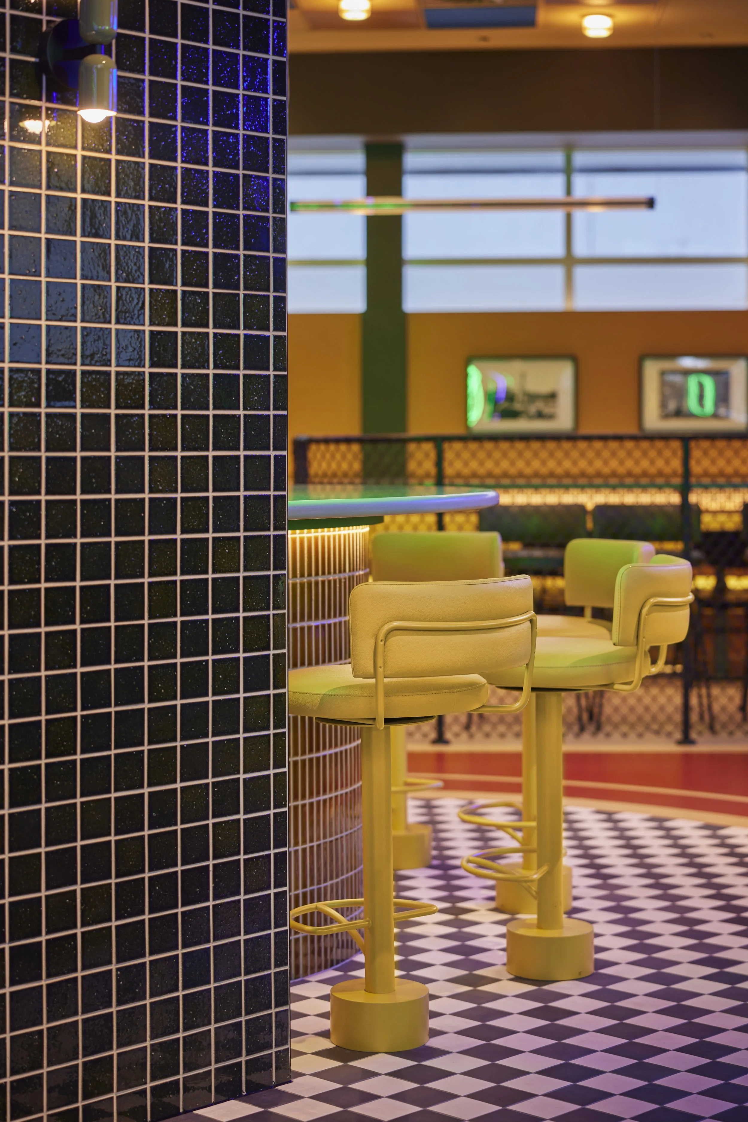 Empty retro-style diner with black tiled walls, yellow bar stools, checkered floor, and neon-lit decor.