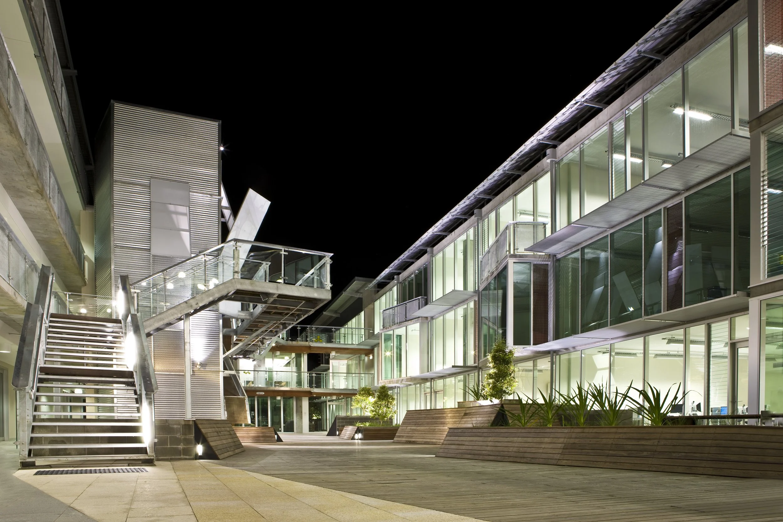 Night view of modern commercial building with glass windows, outdoor stairs, and wooden planters with plants.