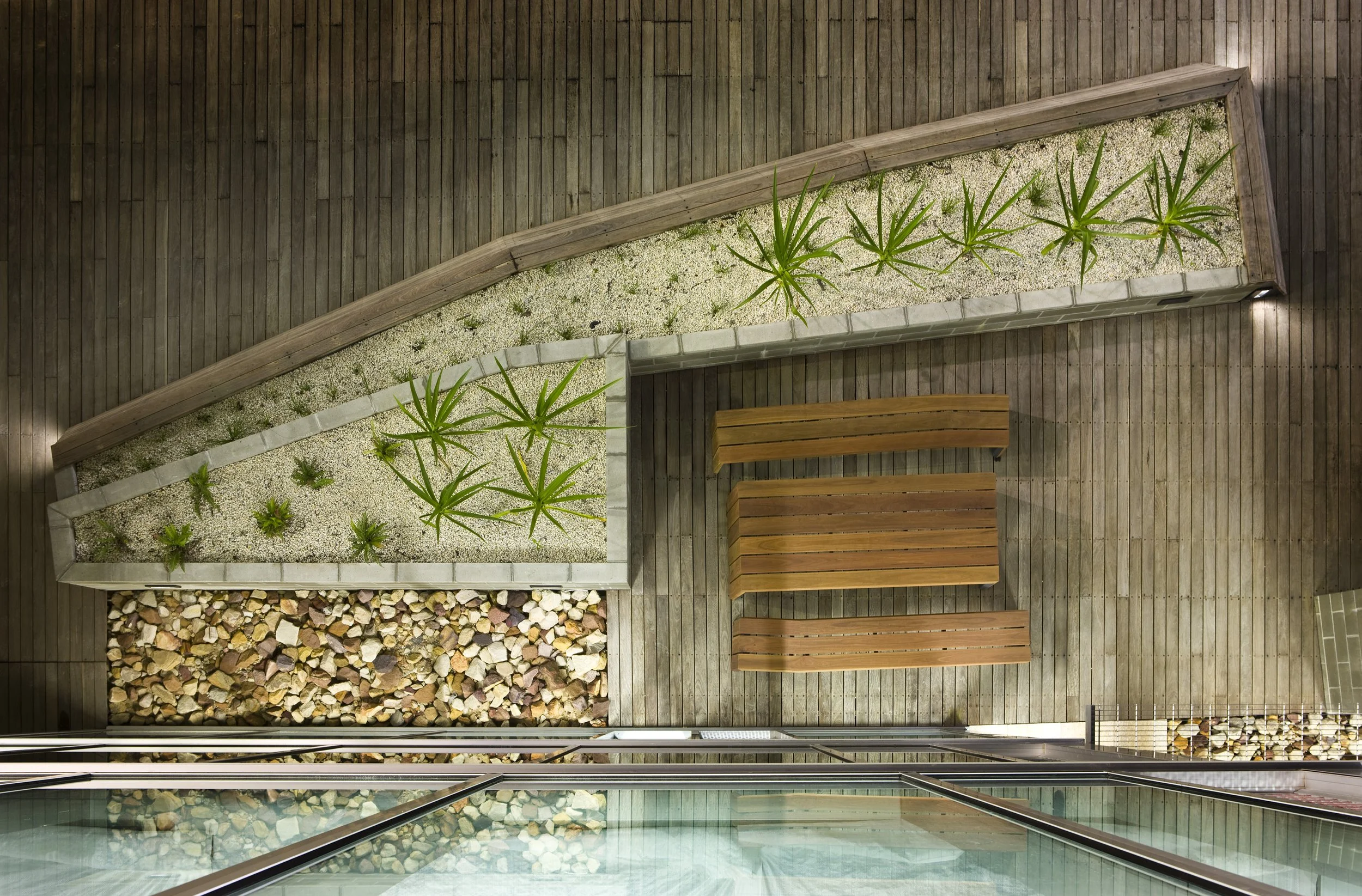 Top-down view of an outdoor balcony with wooden flooring, three wooden benches, a planter with green plants and white gravel, and a wall of glass doors.