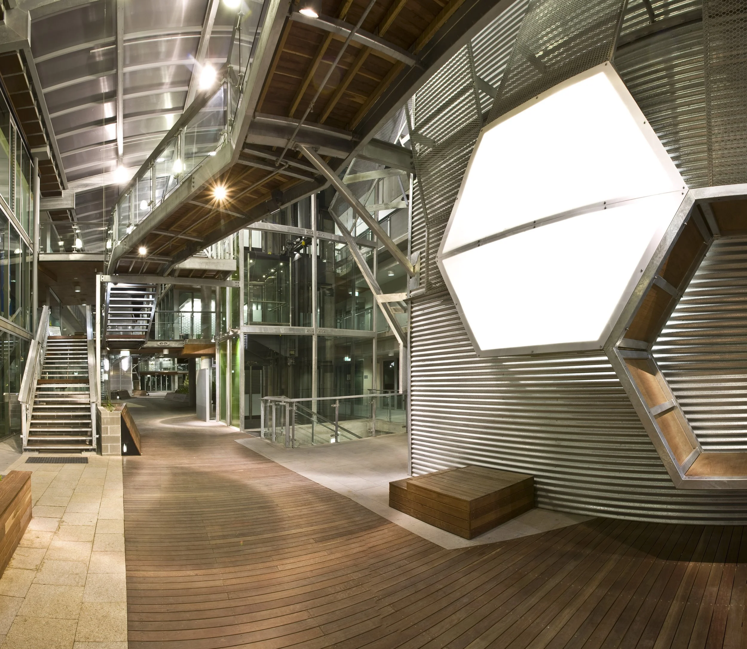 Modern architectural interior with glass walls, metal structures, wooden flooring, stairs, and a large illuminated hexagonal feature on the wall.
