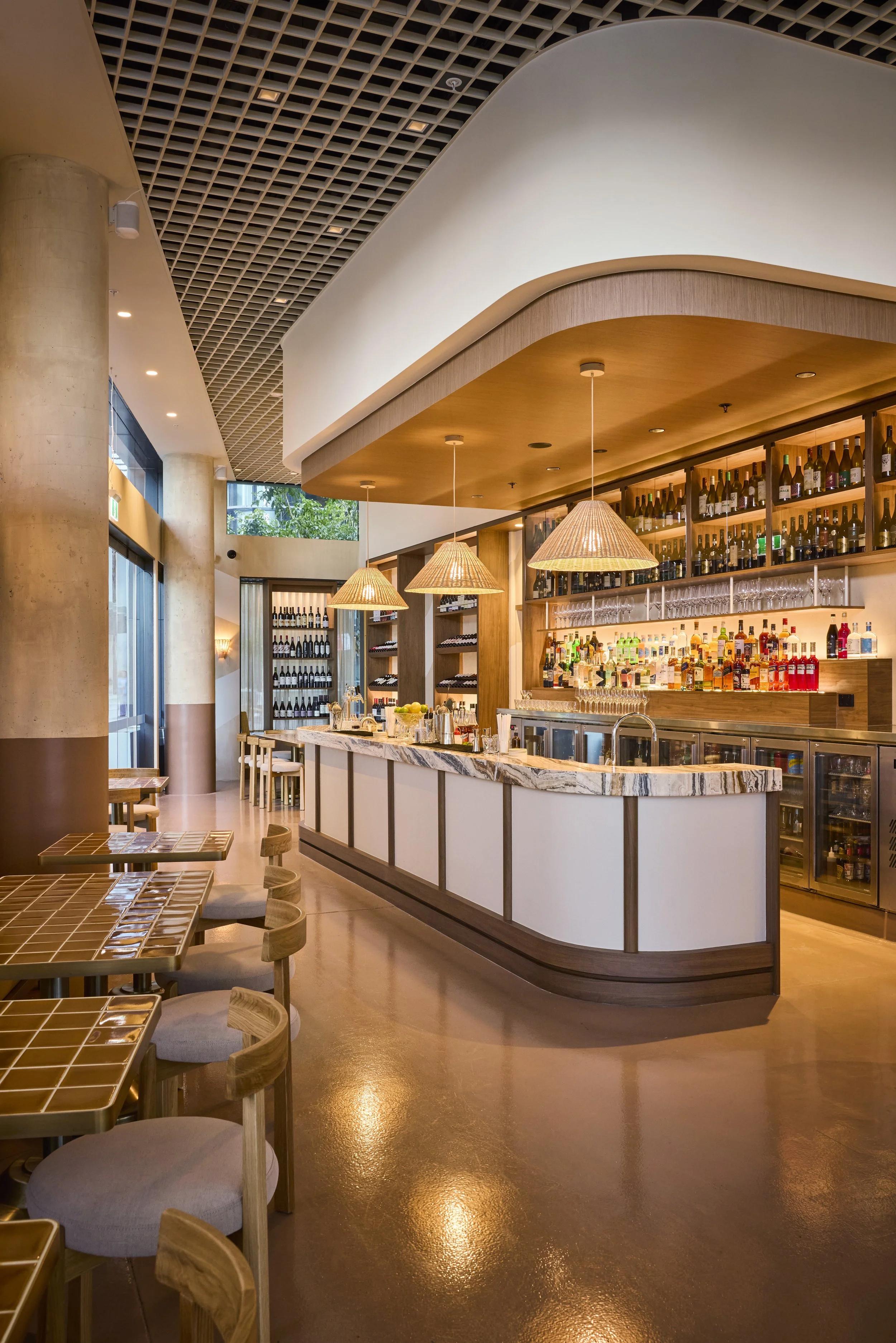 Interior of a modern bar with a white marble counter, wooden shelves stocked with bottles, hanging pendant lights, and seating area with tables and chairs.