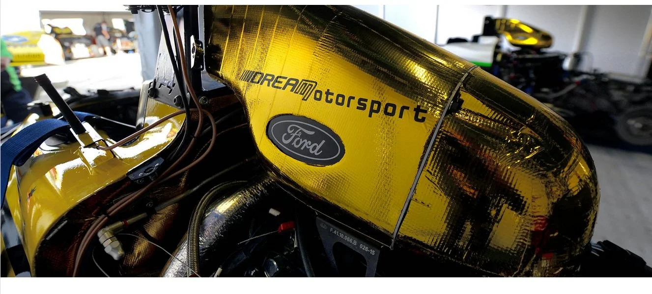 Close-up of a racing car's yellow body with 'DREAM Motorsport' and Ford logos, inside a garage with tools and equipment in the background.