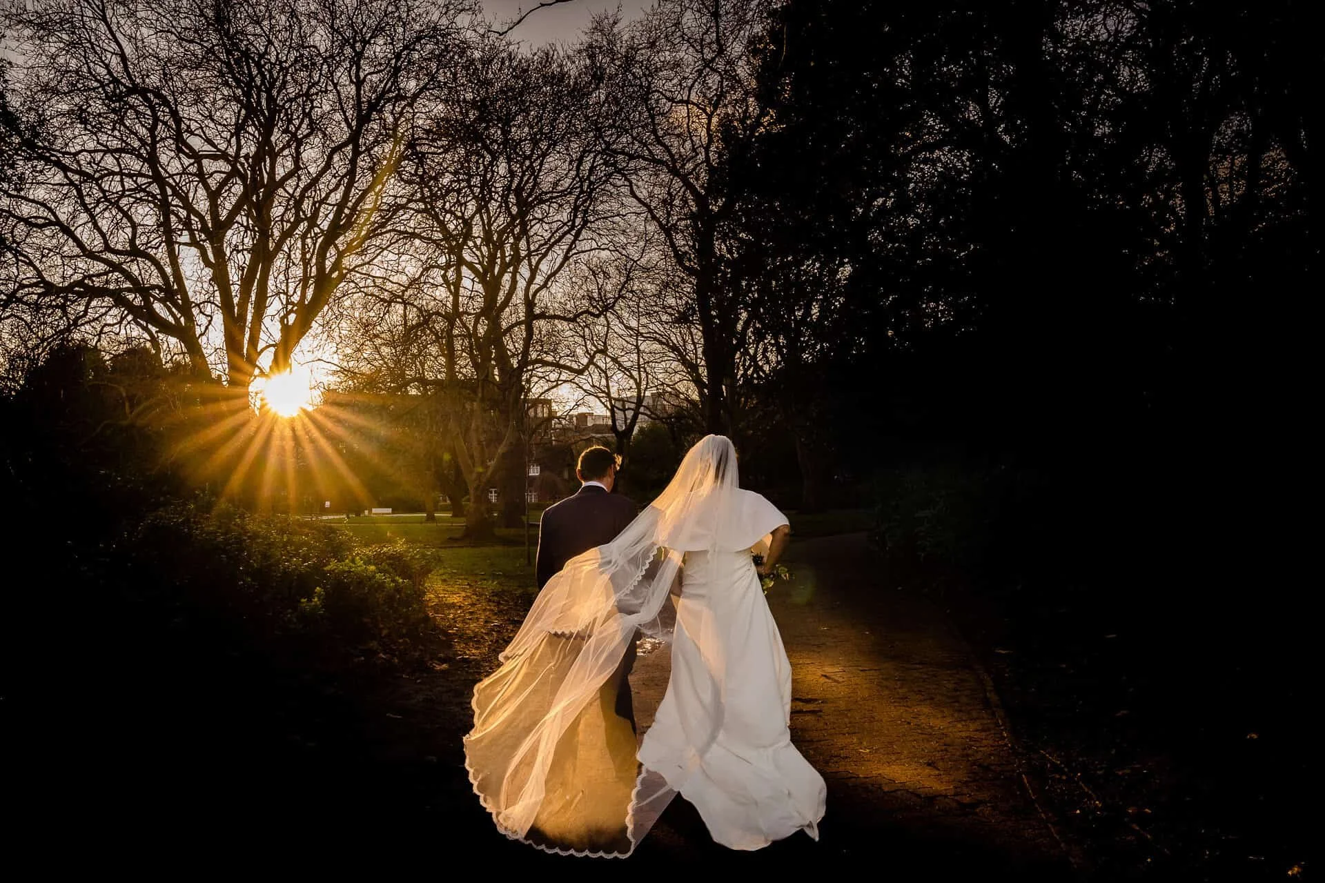Bride walking under trees at sunset, her veil and dress glowing in the low light