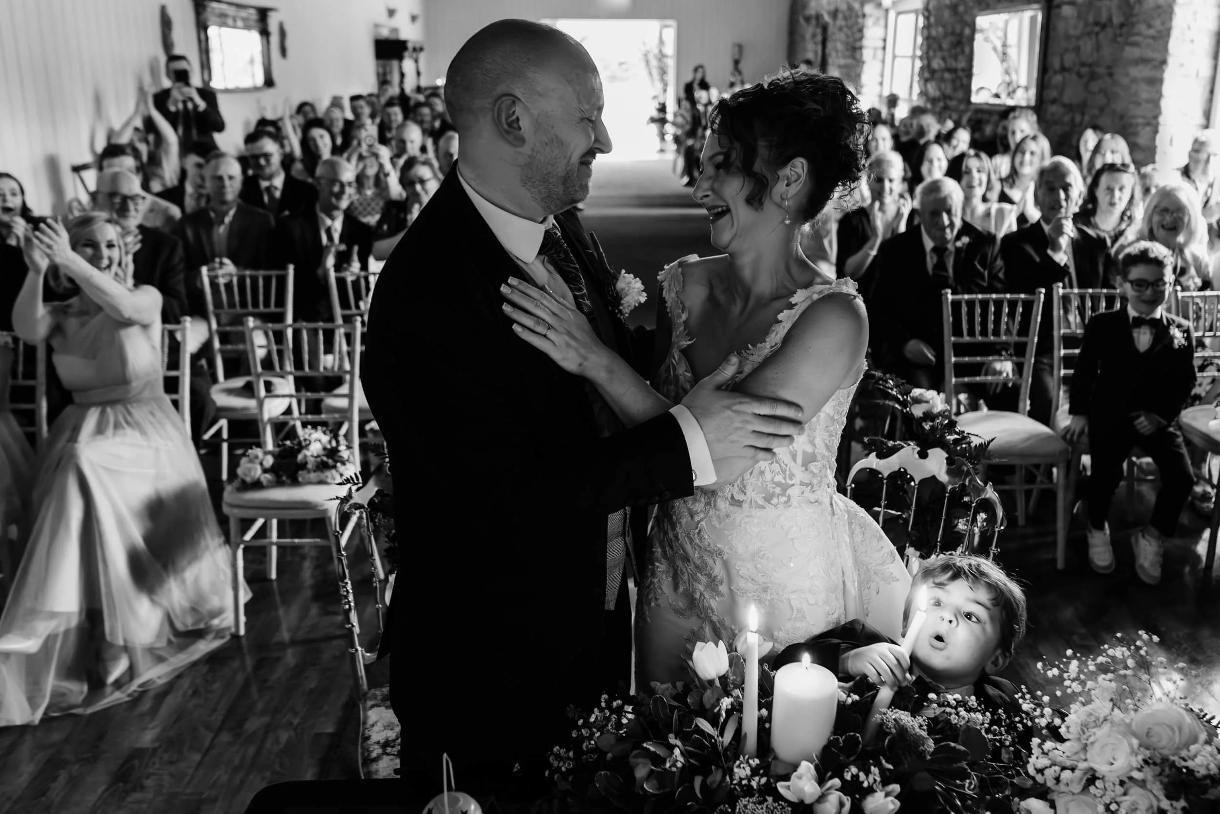 Black and white photograph of a couple embracing at their ceremony while a young guest looks up at them