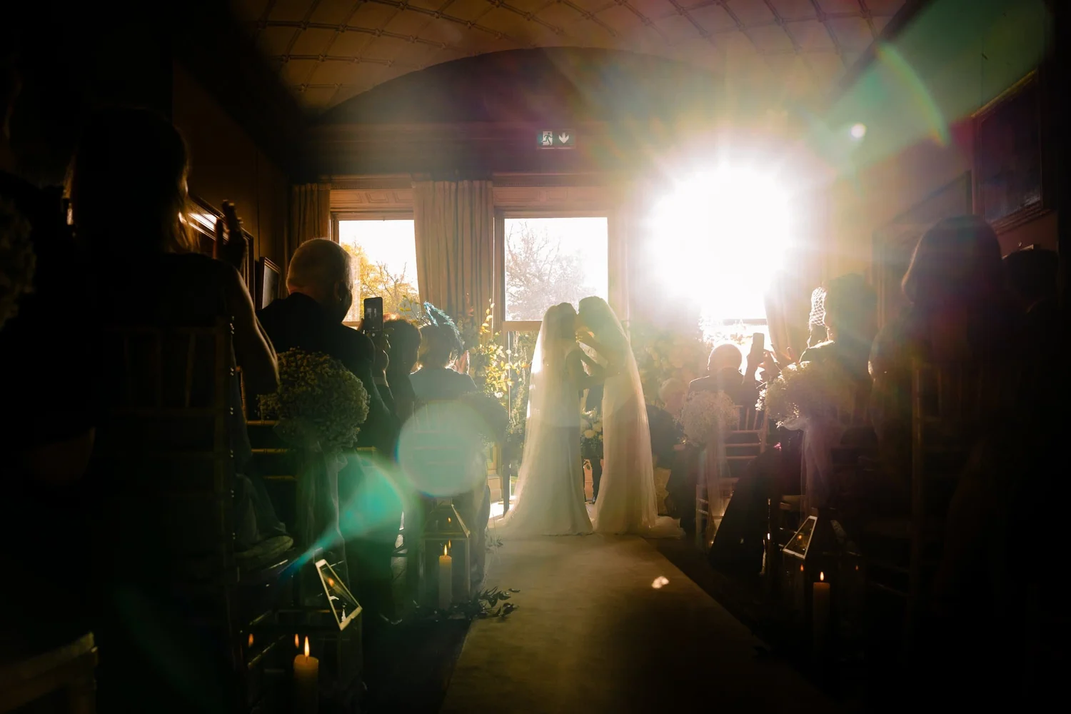 Two brides kissing at the top of the aisle, backlit by low sun streaming through the windows at Castle Leslie