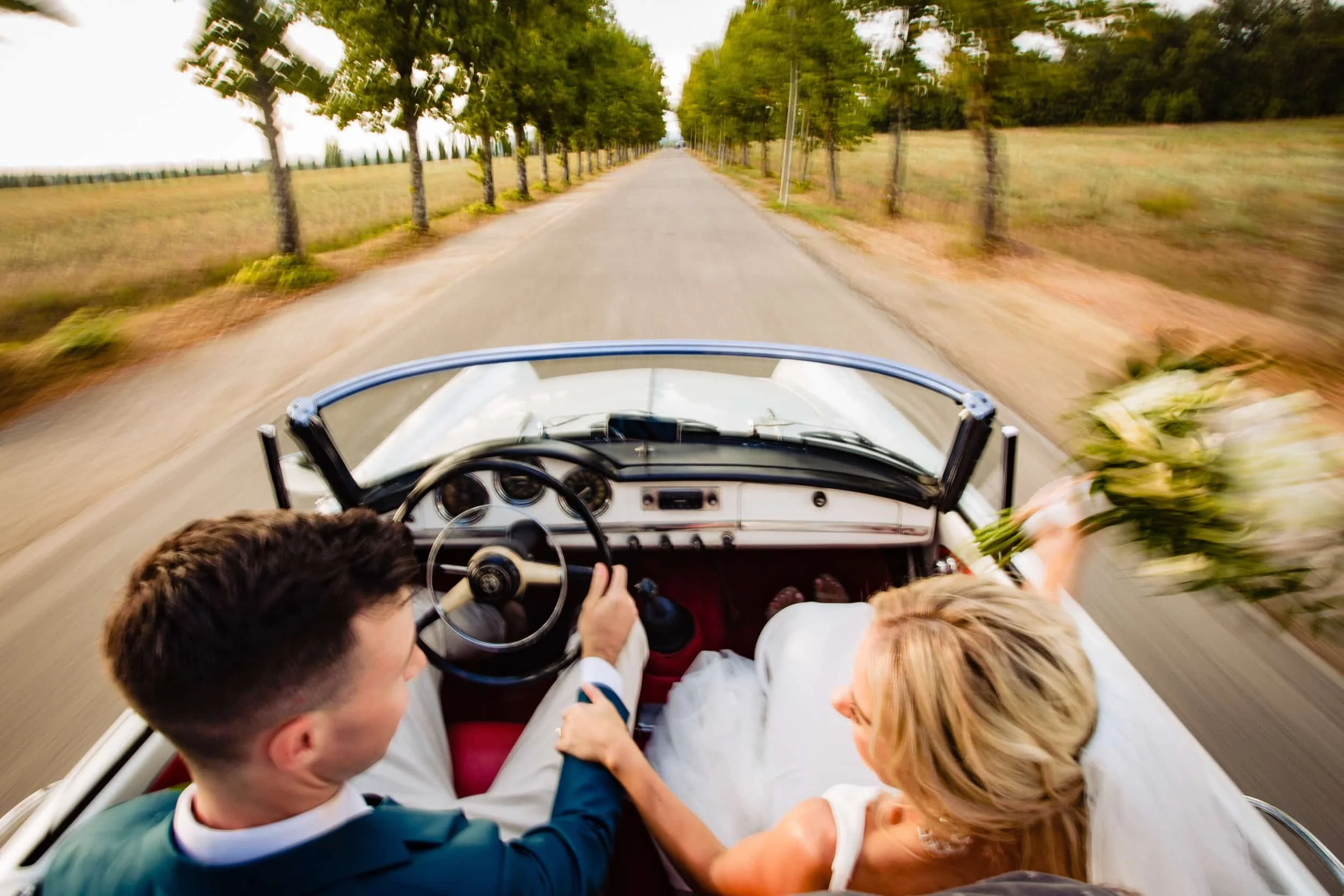 Couple laughing in a vintage convertible on a tree-lined road in Italy