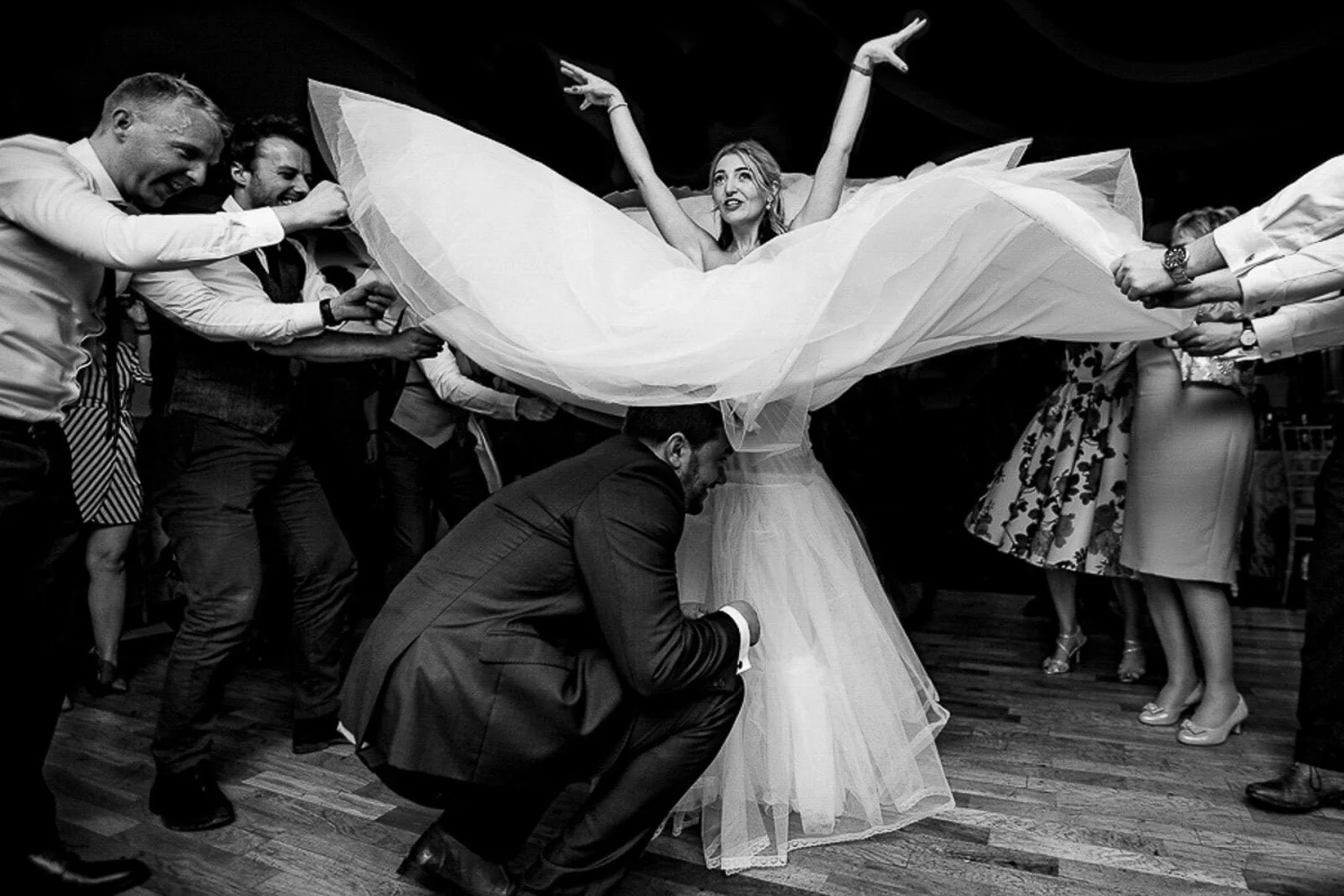 Black and white photograph of a bride's dress billowing as guests celebrate around her on the dance floor