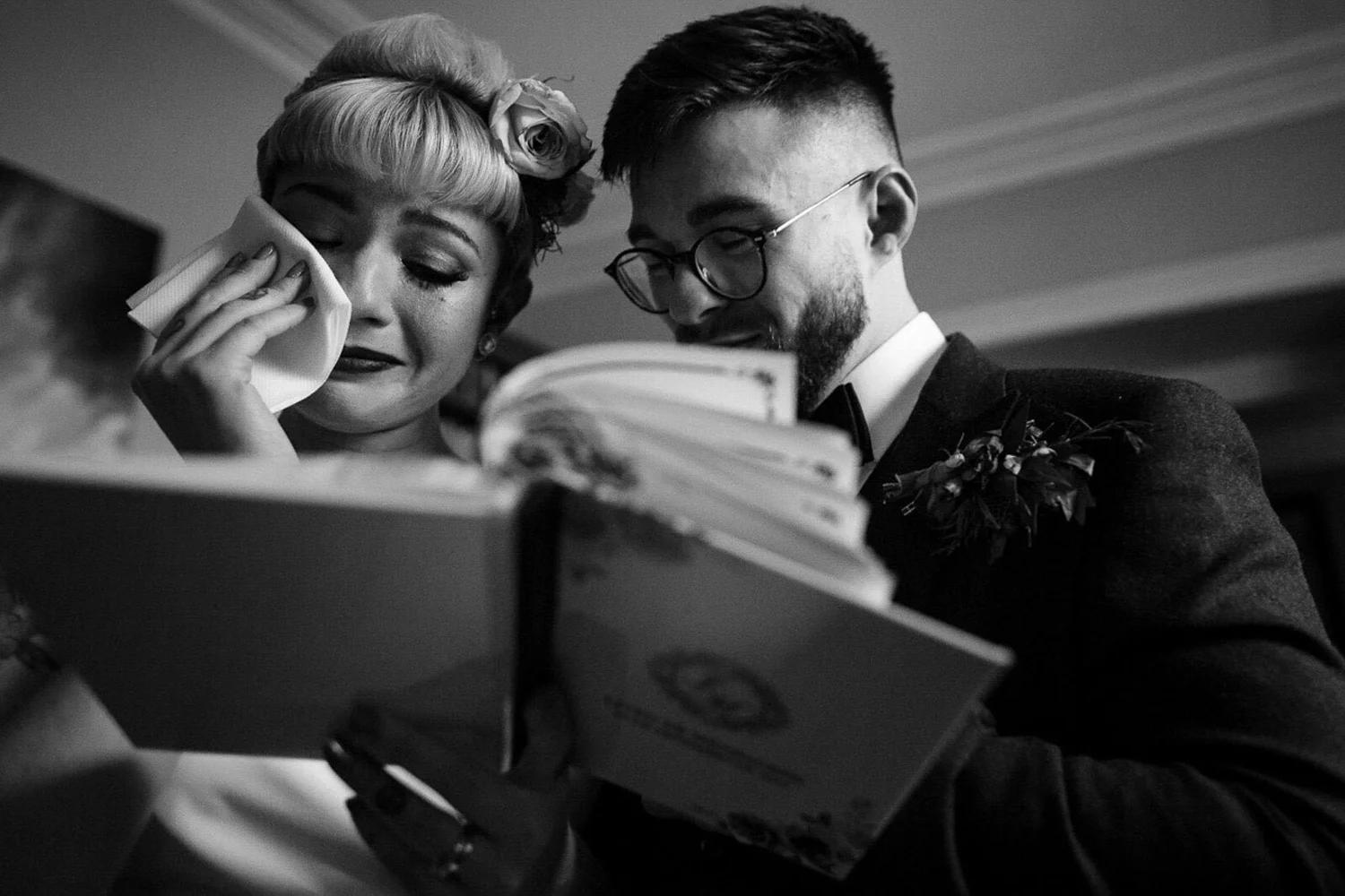 Black and white photograph of a bride wiping away tears beside her partner as they read from the ceremony booklet