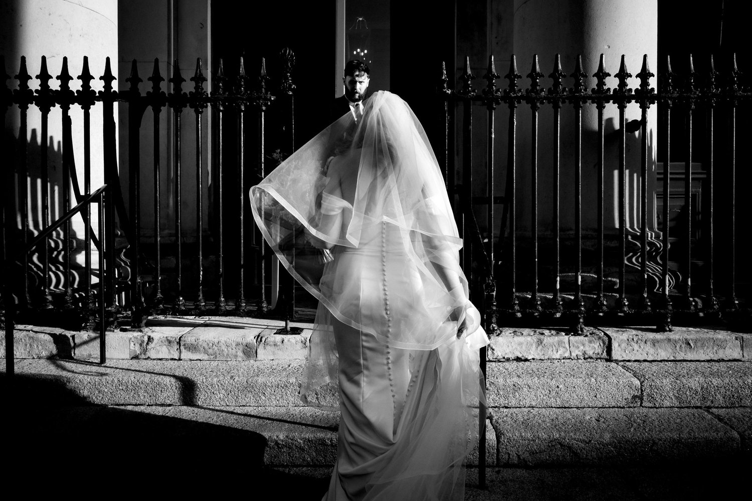 Black and white photograph of a bride walking past railings in Dublin, her veil lifting in the breeze