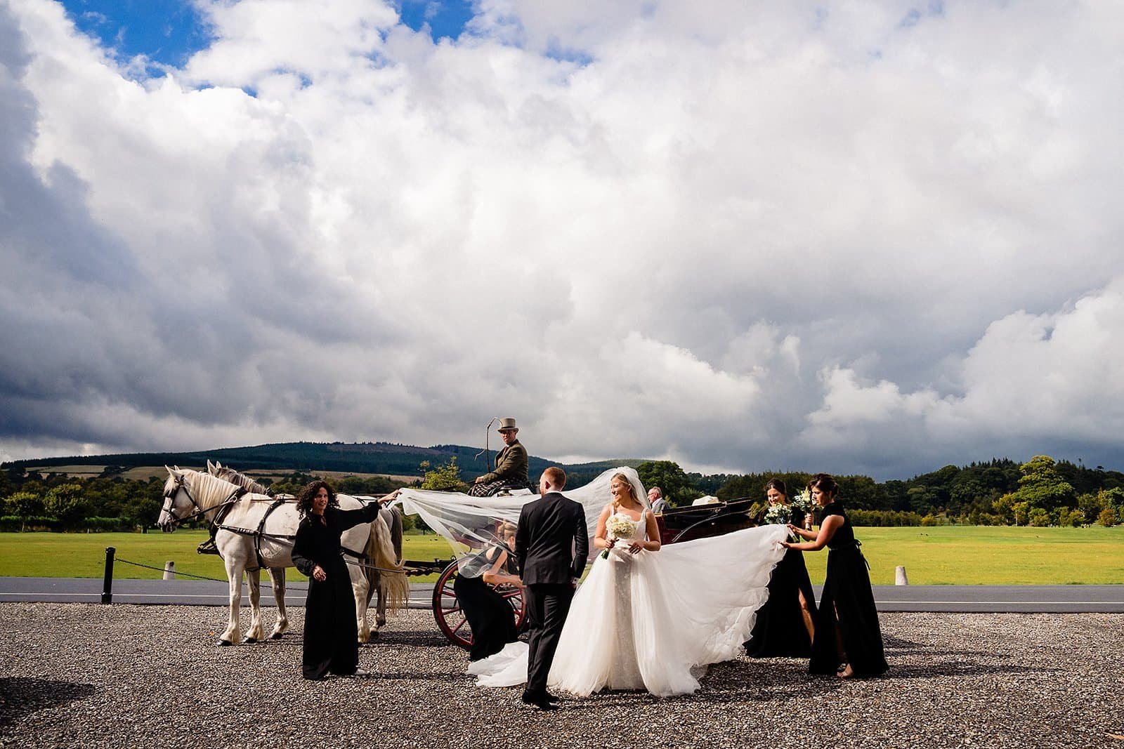 Wedding scene with bride and groom, a horse-drawn carriage, and attendants outdoors on a cloudy day.