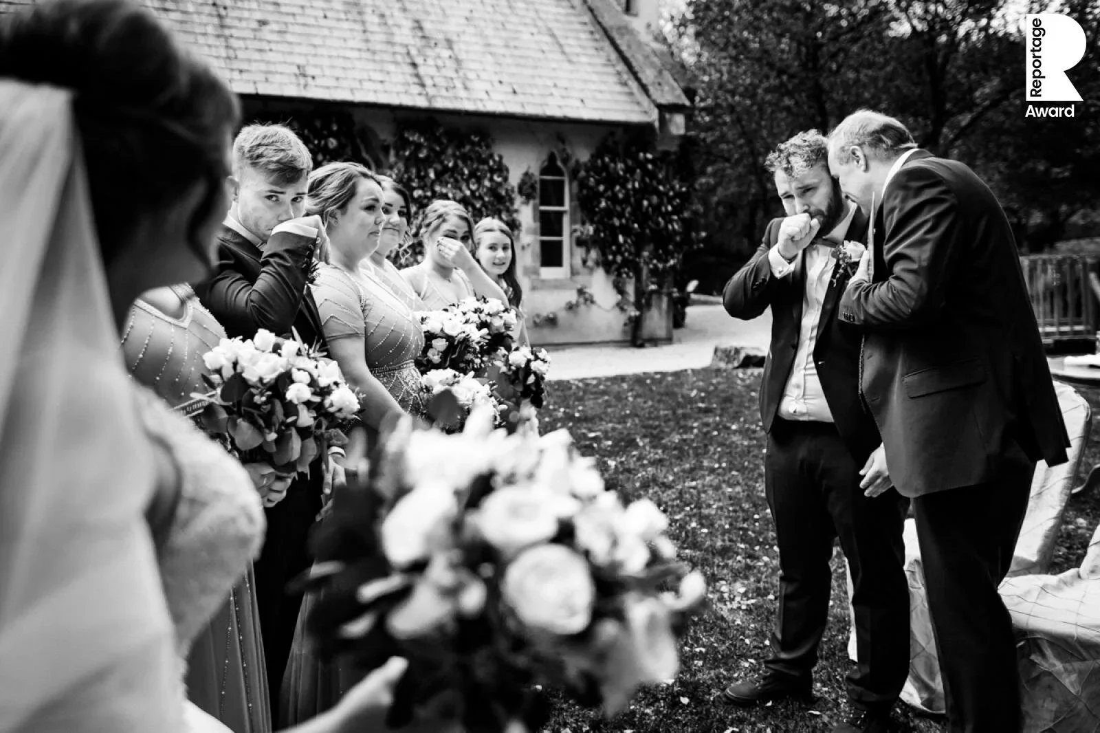 Black and white photo of a wedding ceremony with the bride and groom and wedding party outdoors, with the bride's back to the camera and the groom emotional during vows, surrounded by cheers and emotional participants.