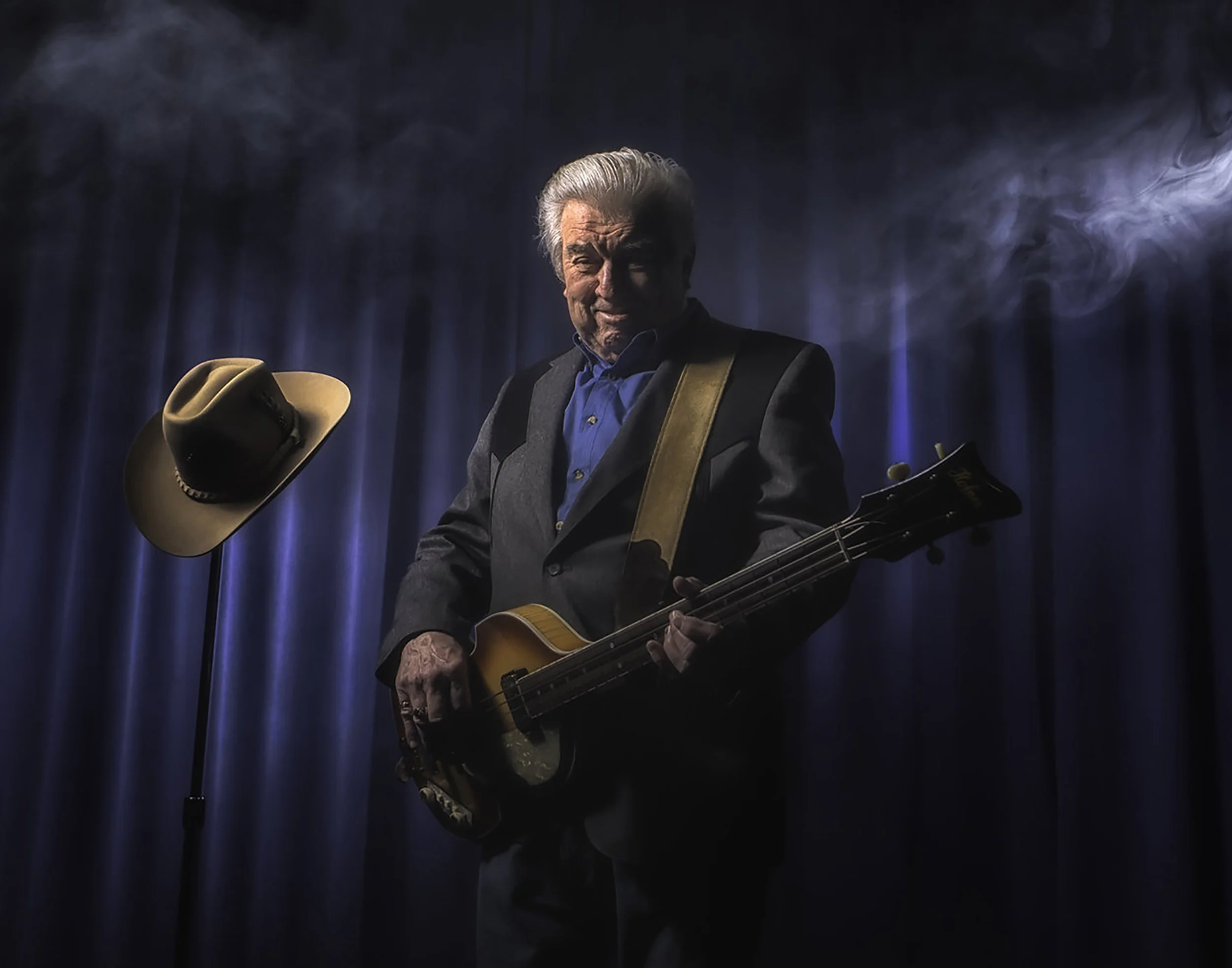 An older man with gray hair playing a guitar on a stage with blue curtains. A cowboy hat hangs on a stand next to him, and there is smoke in the background.