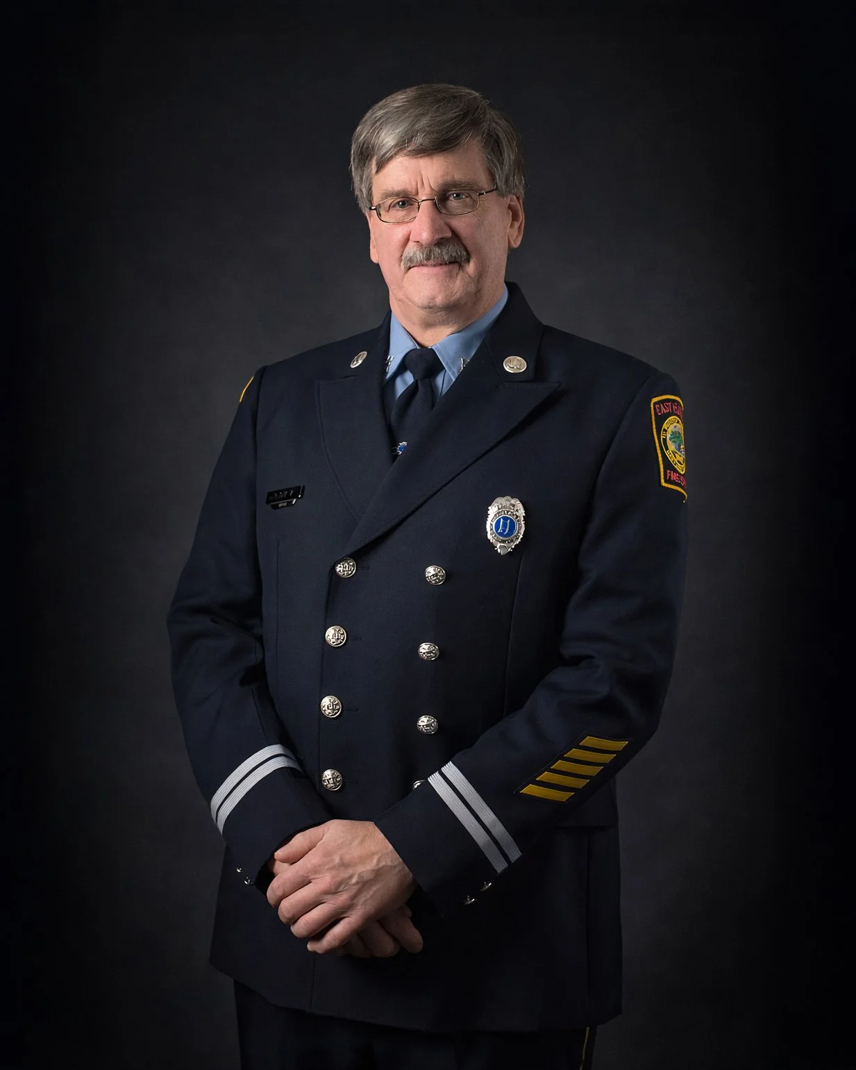 A male firefighter in uniform with a badge, standing against a dark background.