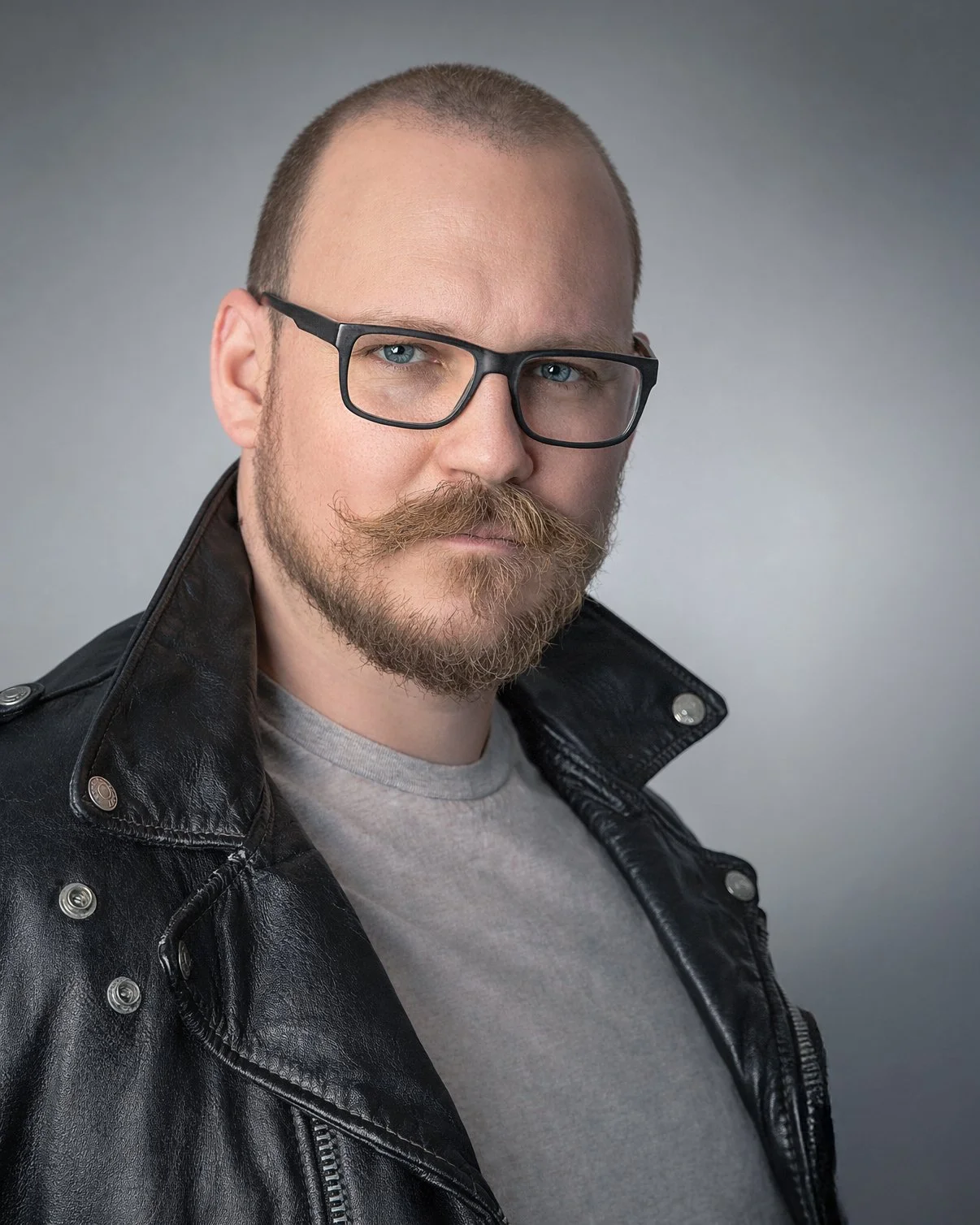 A man with a beard and mustache wearing black glasses, a gray t-shirt, and a black leather jacket. He is looking directly at the camera with a serious expression.