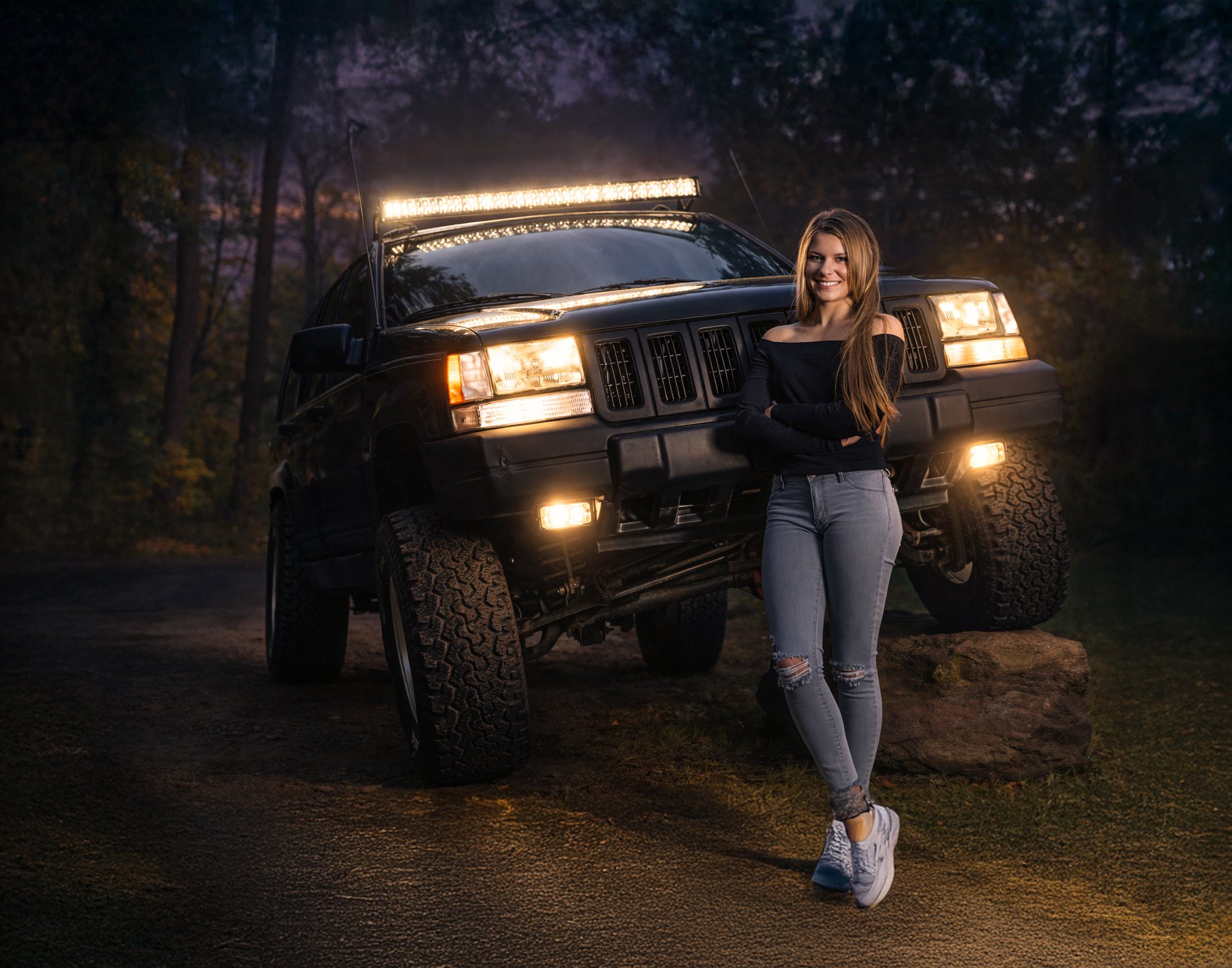 A young woman leaning against a large black SUV with off-road tires during twilight in a wooded area. She is smiling, crossing her arms, wearing a black off-shoulder top, ripped jeans, and sneakers.