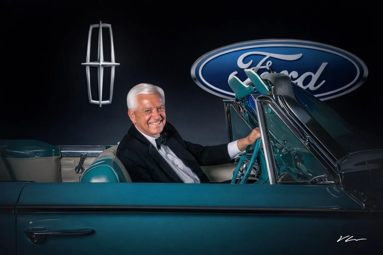 An elderly man with gray hair, dressed in a tuxedo, smiling while sitting in a vintage convertible car with a teal exterior. The background includes a large Ford logo and an iconic Lincoln emblem, indicating a car showroom or event for Ford and Linco
