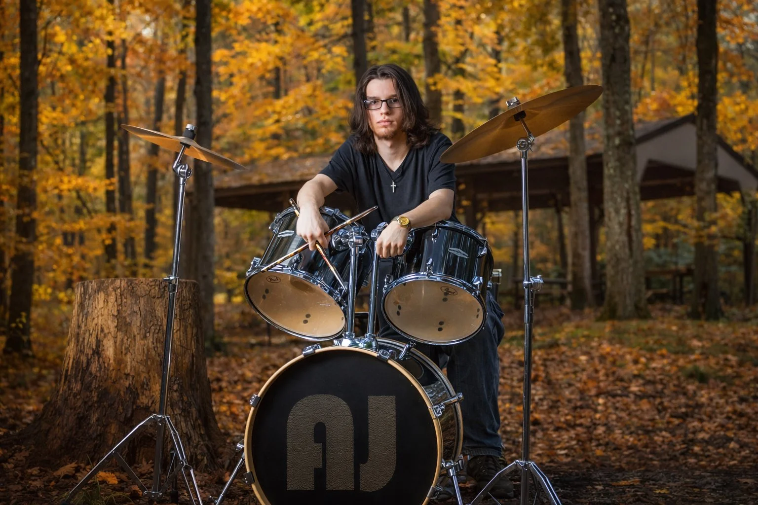 A young man with long dark hair, glasses, and a black t-shirt sitting behind a black drum set with the number 10 on the bass drum in a wooded area during autumn with fallen leaves and trees with orange and yellow leaves.