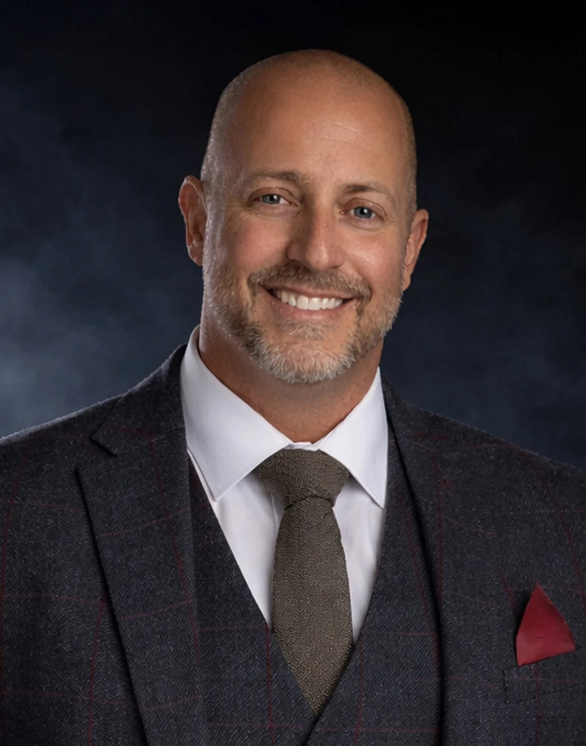 Self Portrait of Vincent Caruso, Close-up portrait of a smiling, bald man with a beard, wearing a dark plaid suit, white shirt, and brown tie, with a dark background.