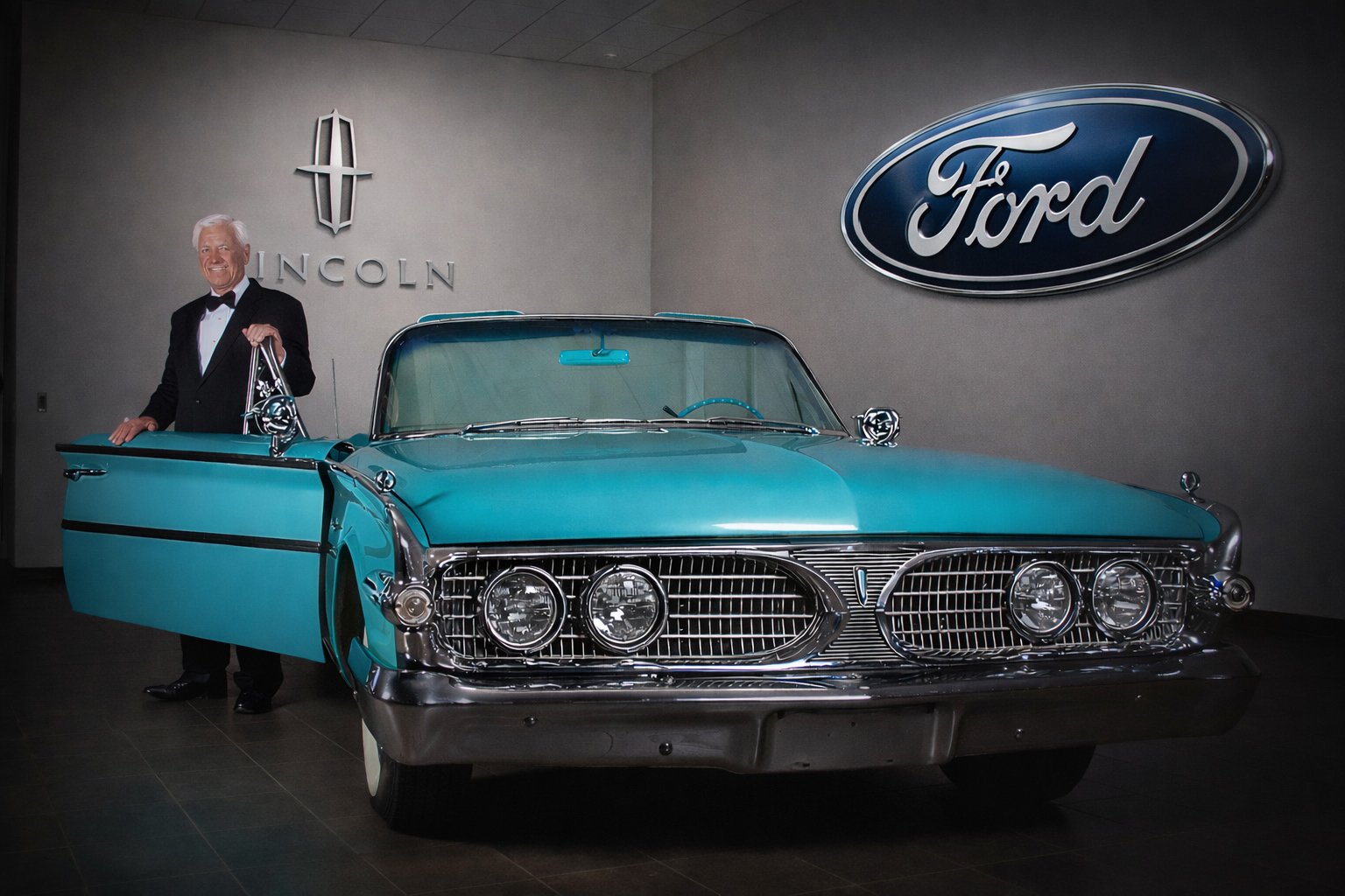 A vintage blue Lincoln car with a man in a tuxedo standing beside it inside a showroom. The background features Lincoln and Ford logos on the wall.