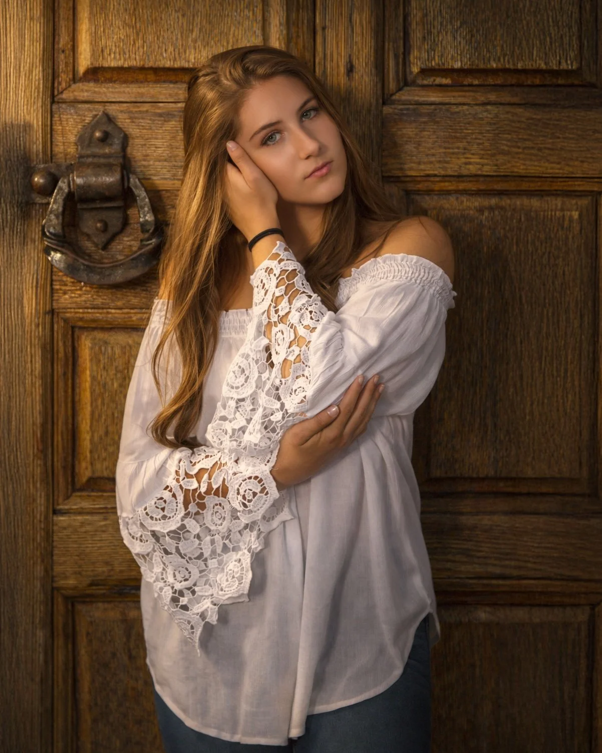 A young woman with long, wavy brown hair, wearing a white off-the-shoulder blouse with intricate lace detailing on the sleeves, standing in front of a wooden door with a decorative metal door knocker. She has a neutral expression and her right hand i