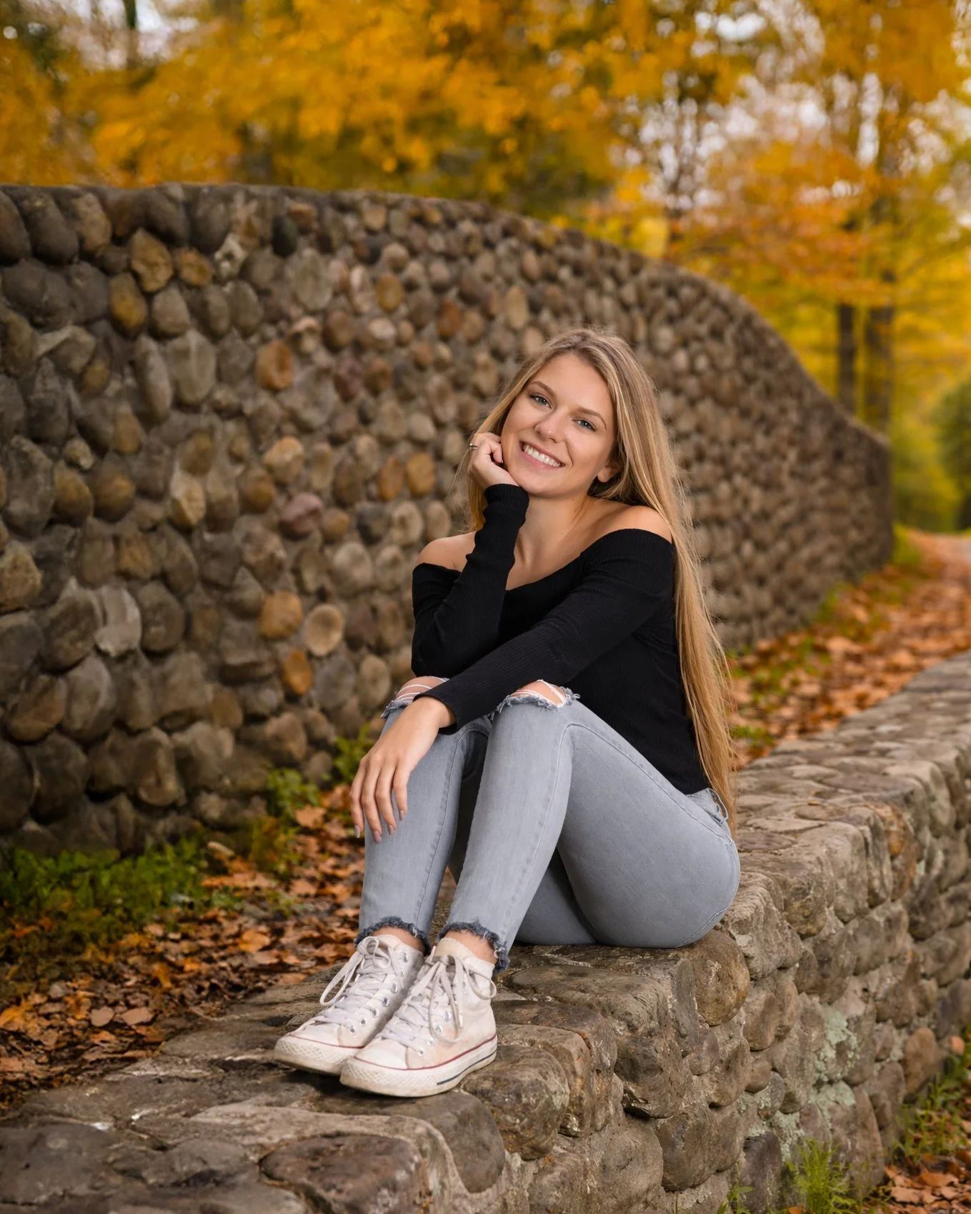 Smiling young woman with long blonde hair sitting on a stone wall in autumn, wearing a black off-the-shoulder sweater, ripped light gray jeans, and white sneakers, with fall foliage behind her.