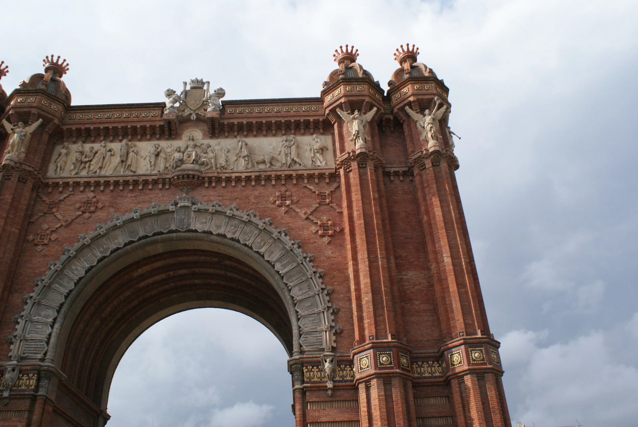 The archway of the Arc de Triumph stands out against the empty park, cement walkways leading up to its brick facade. The details stand triumphant while people mill around its base, looking up.