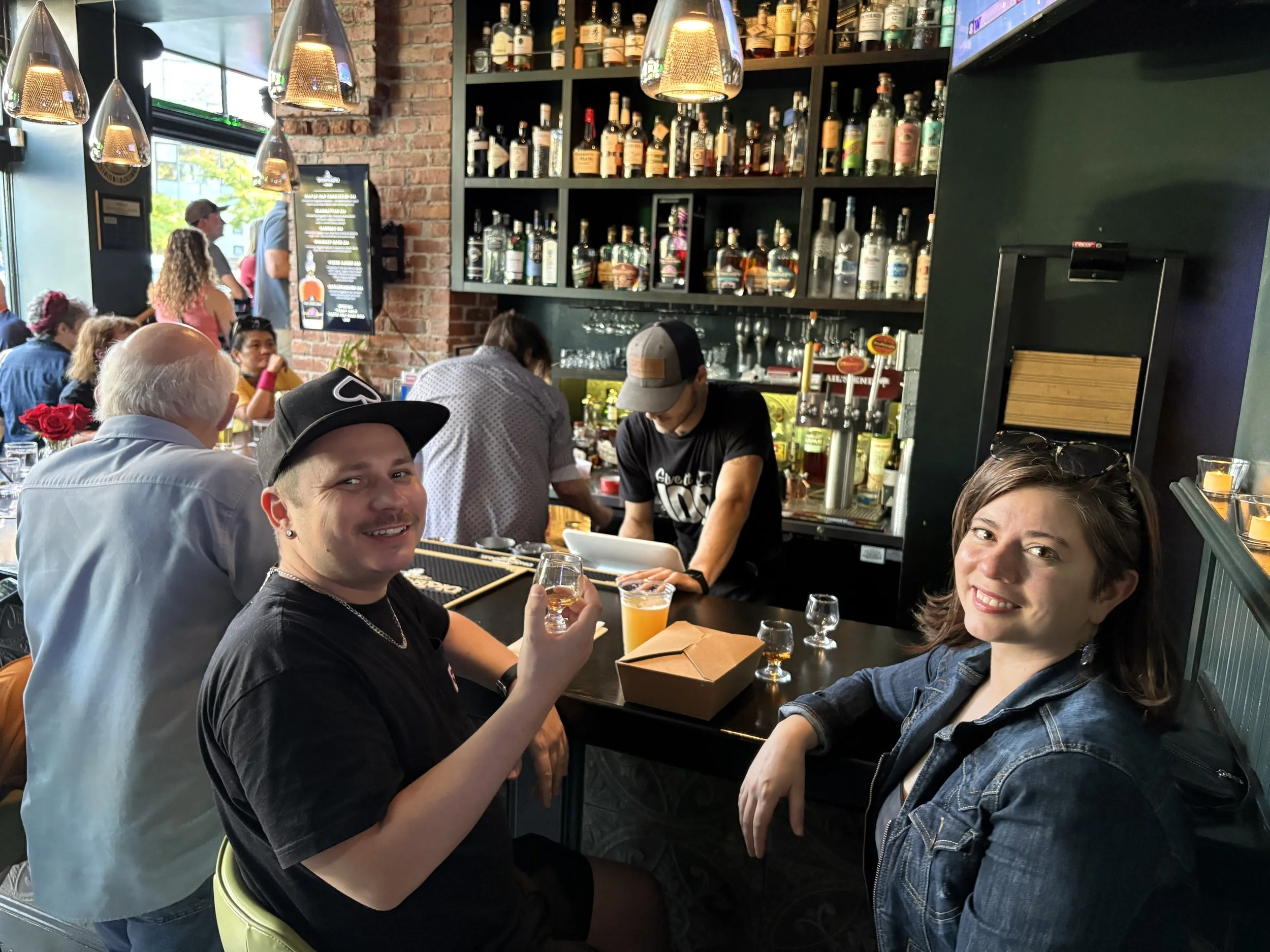 Guests inside The BottleNeck Lounge enjoying a drink at the bar and smiling at the camera with activity going on around them.