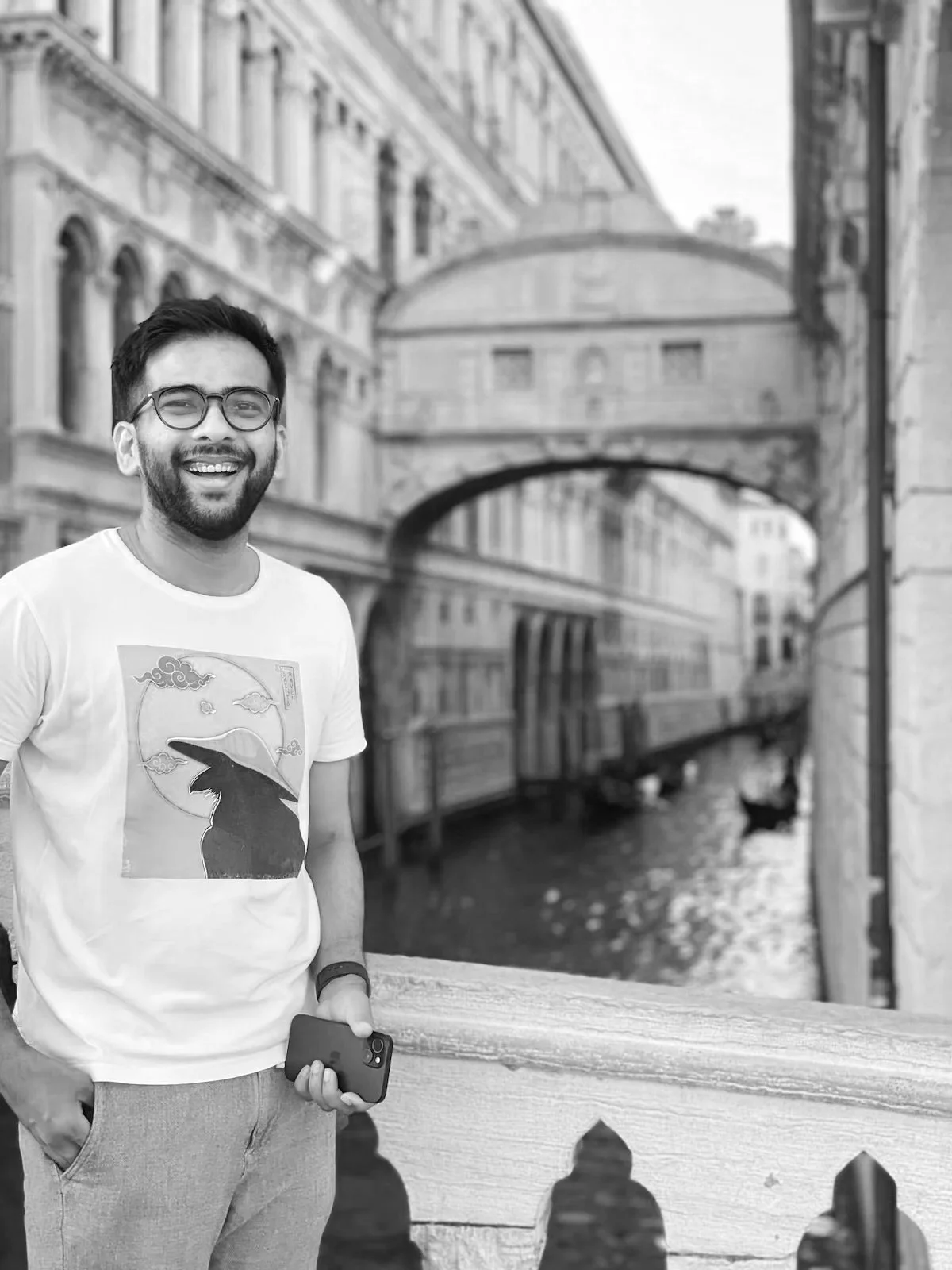 A smiling man with glasses and a beard holding a phone, standing by a bridge with historic buildings and water below in the background.