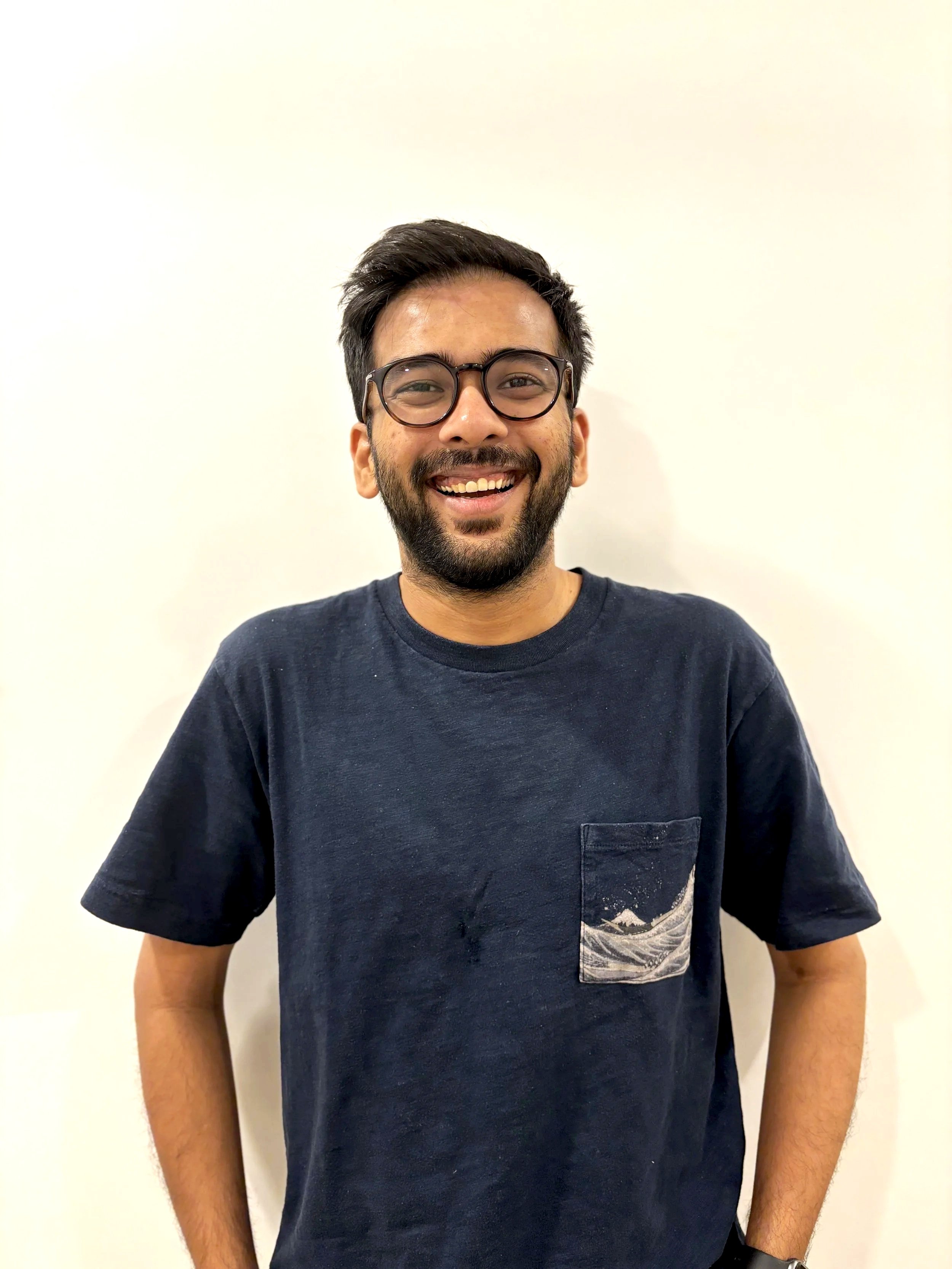 A smiling man with dark hair, glasses, and a beard wearing a navy t-shirt with a pocket, standing against a plain white wall.