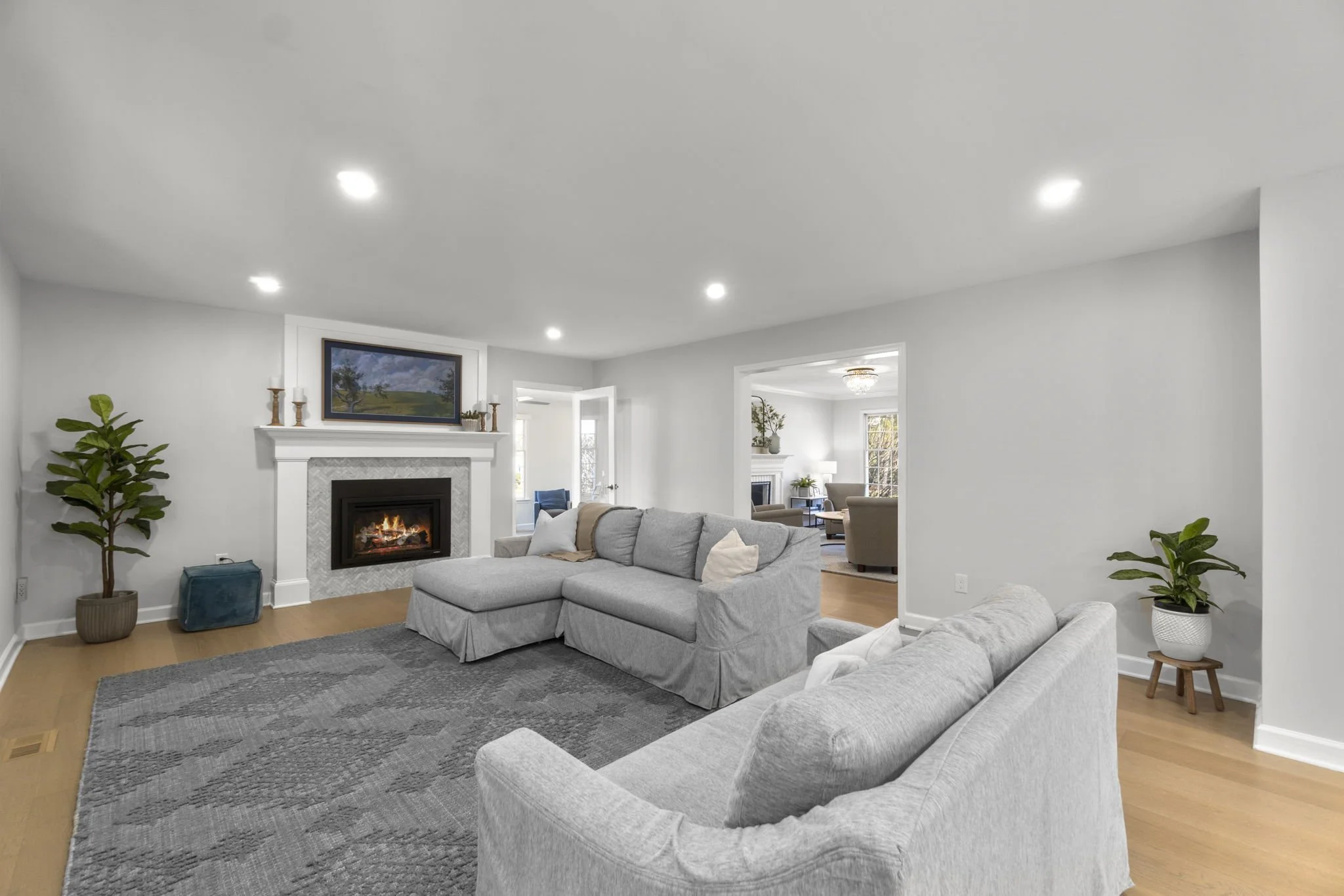 Staged living room with gray sectional sofa, tiled fireplace, area rug, and light wood flooring, opening into adjacent living space