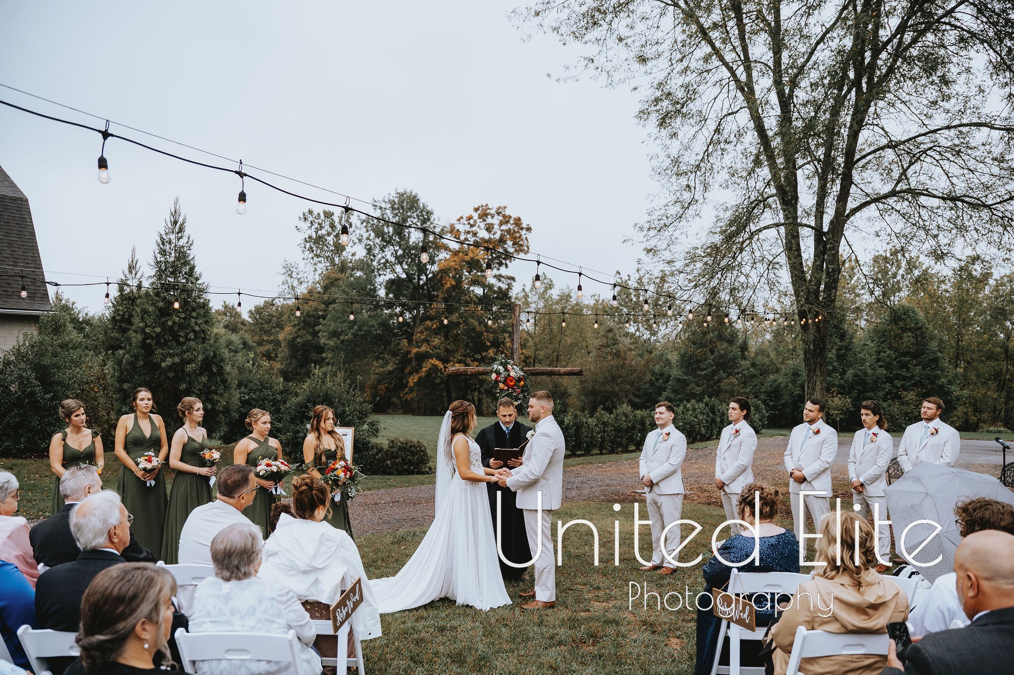 A group of people participating in an outdoor wedding ceremony in a garden setting, with trees and string lights overhead, during daytime.