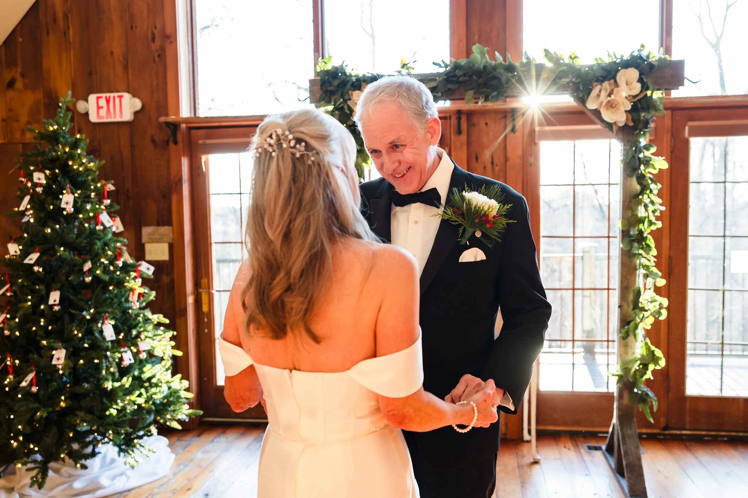 An elderly couple dancing at a wedding reception, with a decorated Christmas tree and a floral arch in the background.