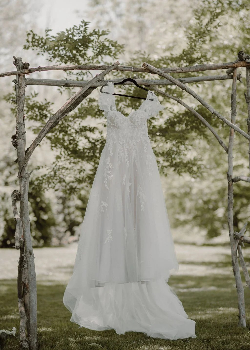 White wedding dress hanging on a black hanger on a rustic wooden frame outdoors, with green foliage in the background.