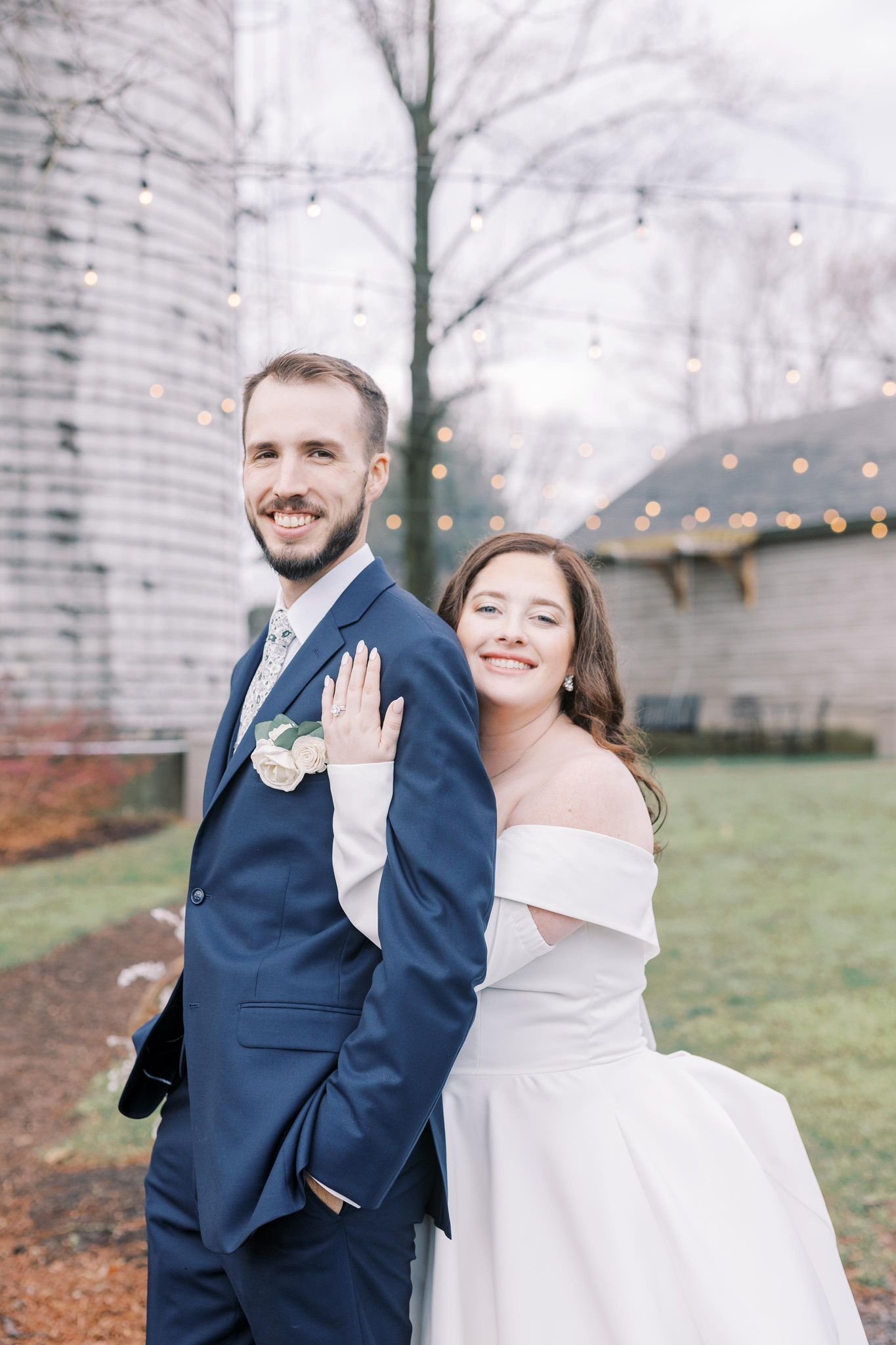 A newlywed couple smiling, with the groom in a navy suit and the bride in a white wedding dress, outdoor garden setting with string lights and trees in the background.