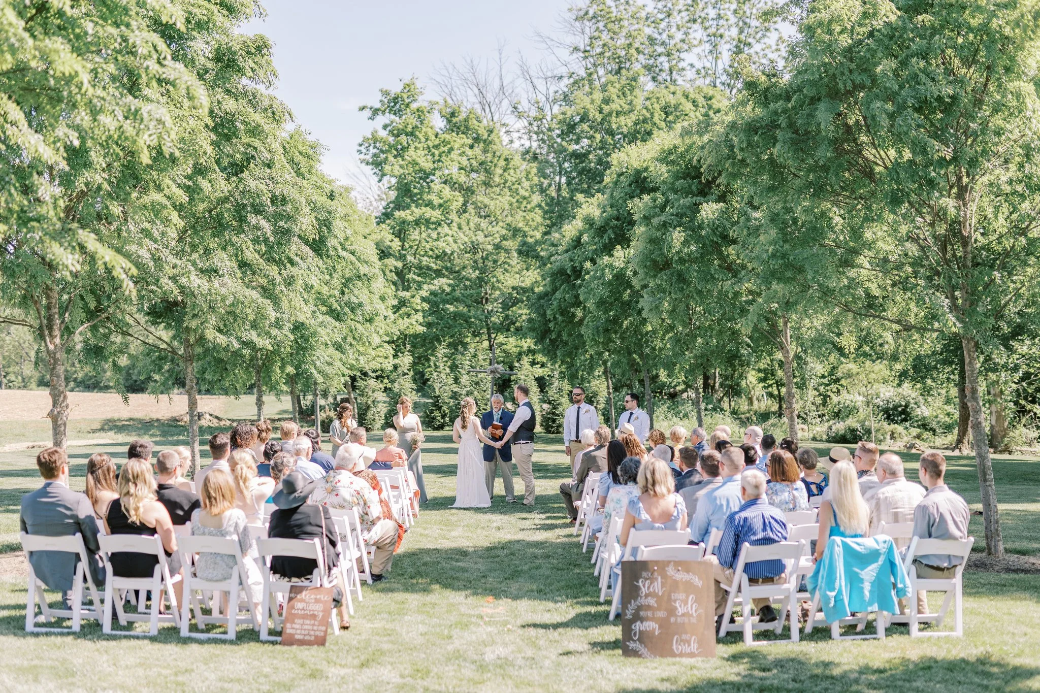 Outdoor wedding ceremony with guests seated on white chairs under green trees, officiant and couple exchanging vows in front of a wooded backdrop.
