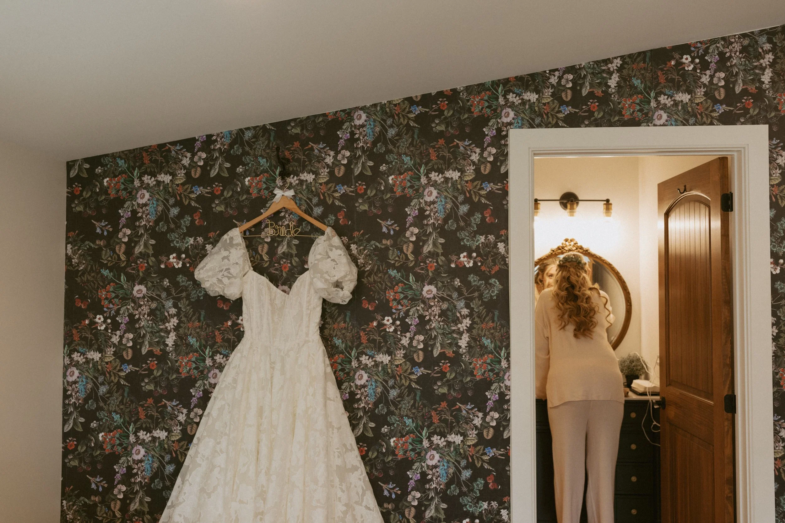 A wedding dress hanging on a floral wallpapered wall in a room, with a woman looking into a mirror in an adjacent room.