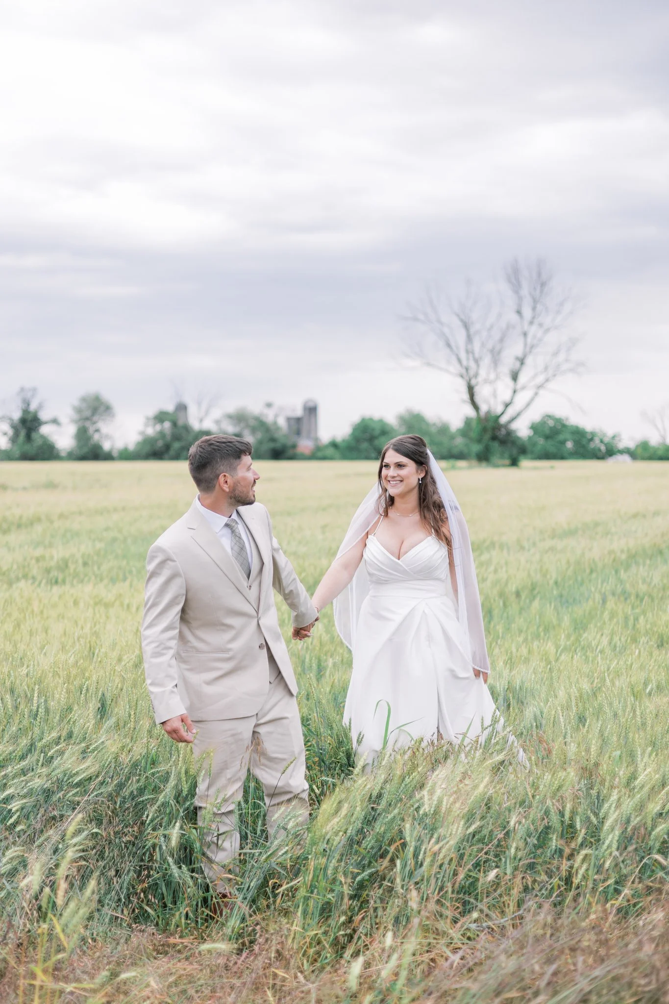A couple dressed in wedding attire standing in a grassy field holding hands and smiling at each other, with trees and a cloudy sky in the background.