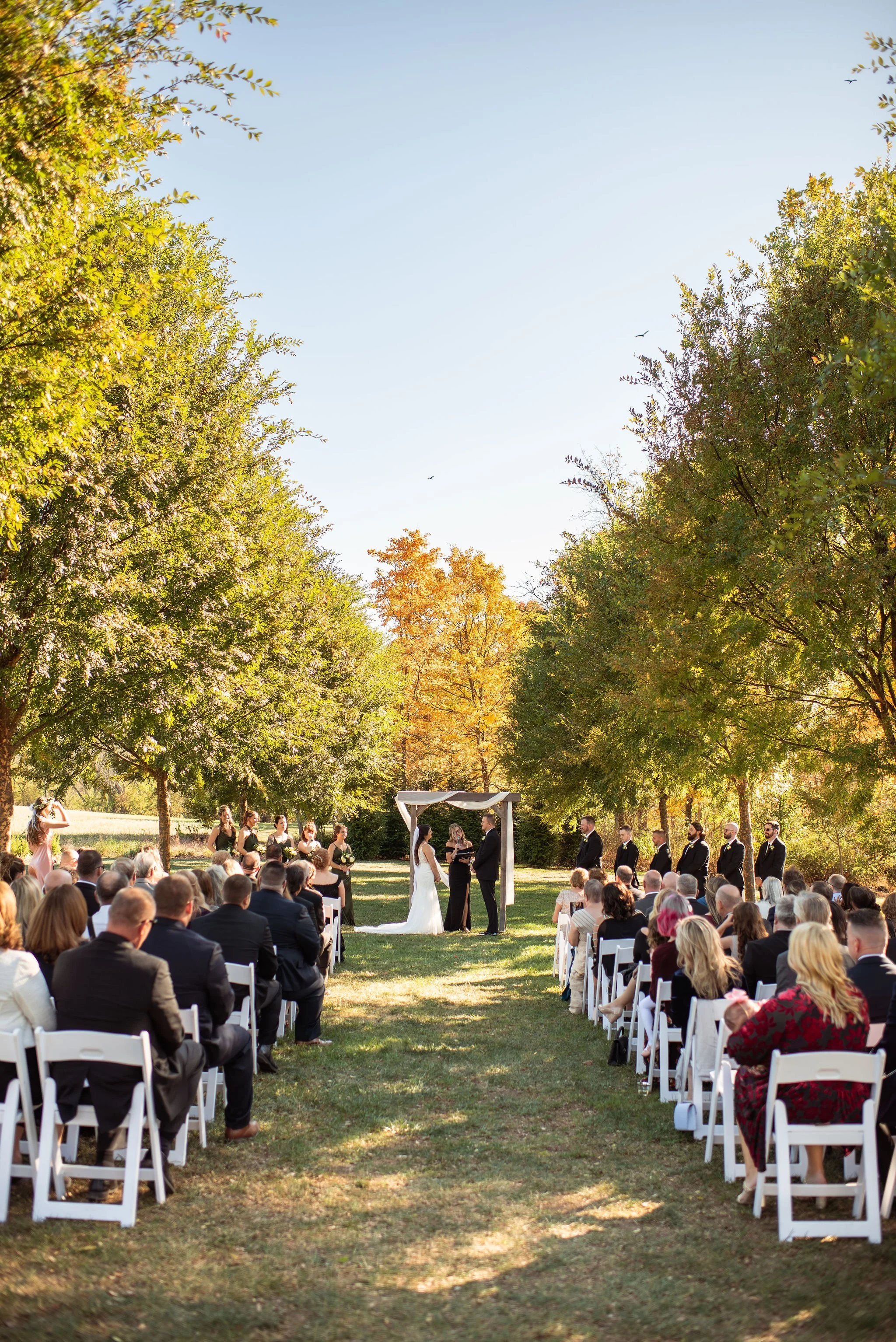 An outdoor wedding ceremony taking place in a park on a sunny day, with guests seated on white chairs facing a small altar where the couple and officiant are standing, surrounded by trees with green and some autumn-colored leaves.