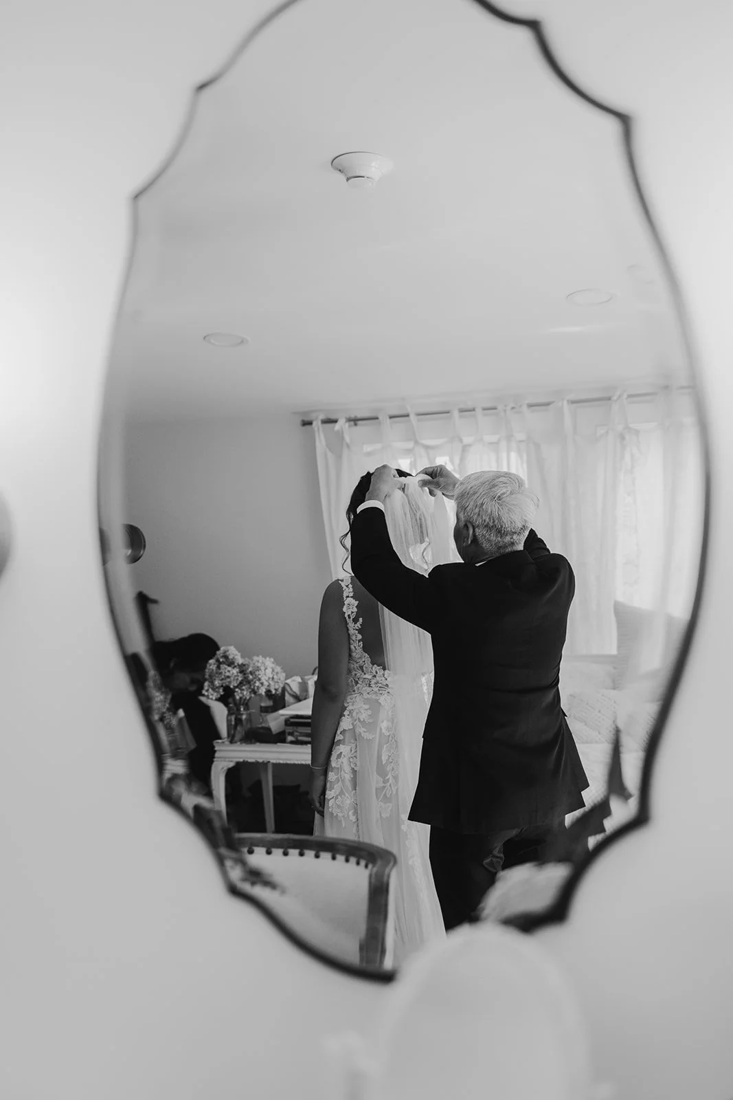 A reflection in a decorative mirror shows a bride and an older man, likely her father, helping her with her veil before her wedding.